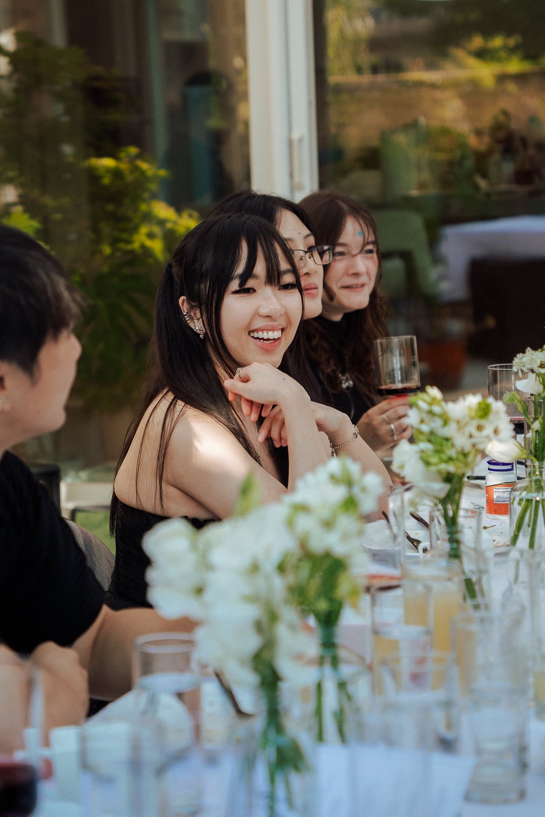 Group of people sitting at a table decorated with white flowers, smiling and enjoying drinks during a social gathering.