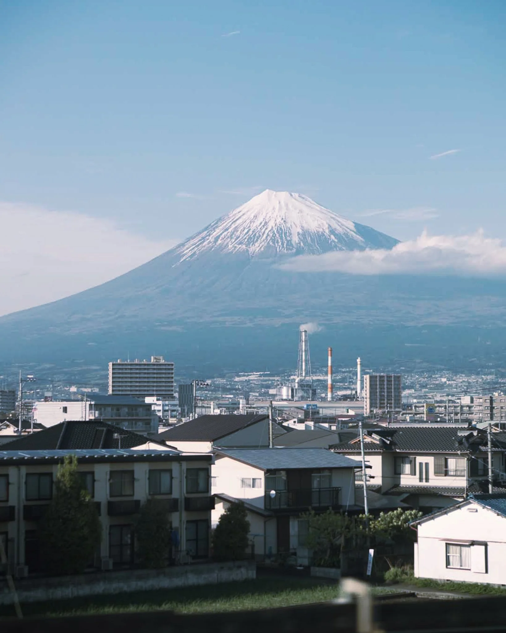 A view of Mount Fuji with snow on its peak, overlooking a city with residential houses and industrial buildings.