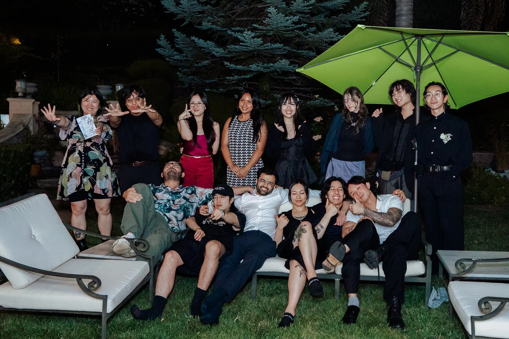 A group of people gathered outdoors at night, sitting and standing on a lawn with patio furniture and a large green umbrella, celebrating together.