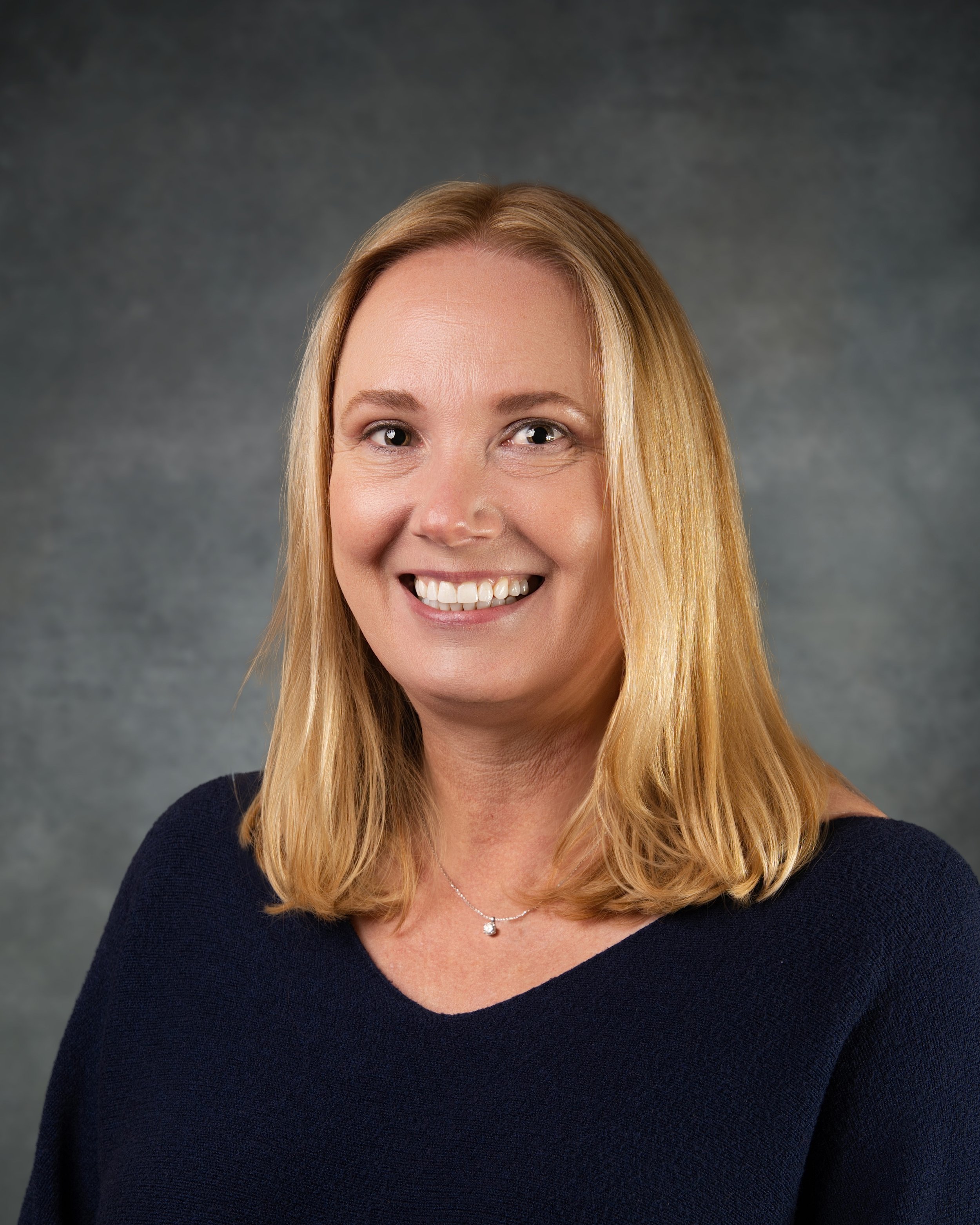 A smiling woman with shoulder-length blonde hair wearing a navy blue top and a delicate necklace, against a textured gray background.