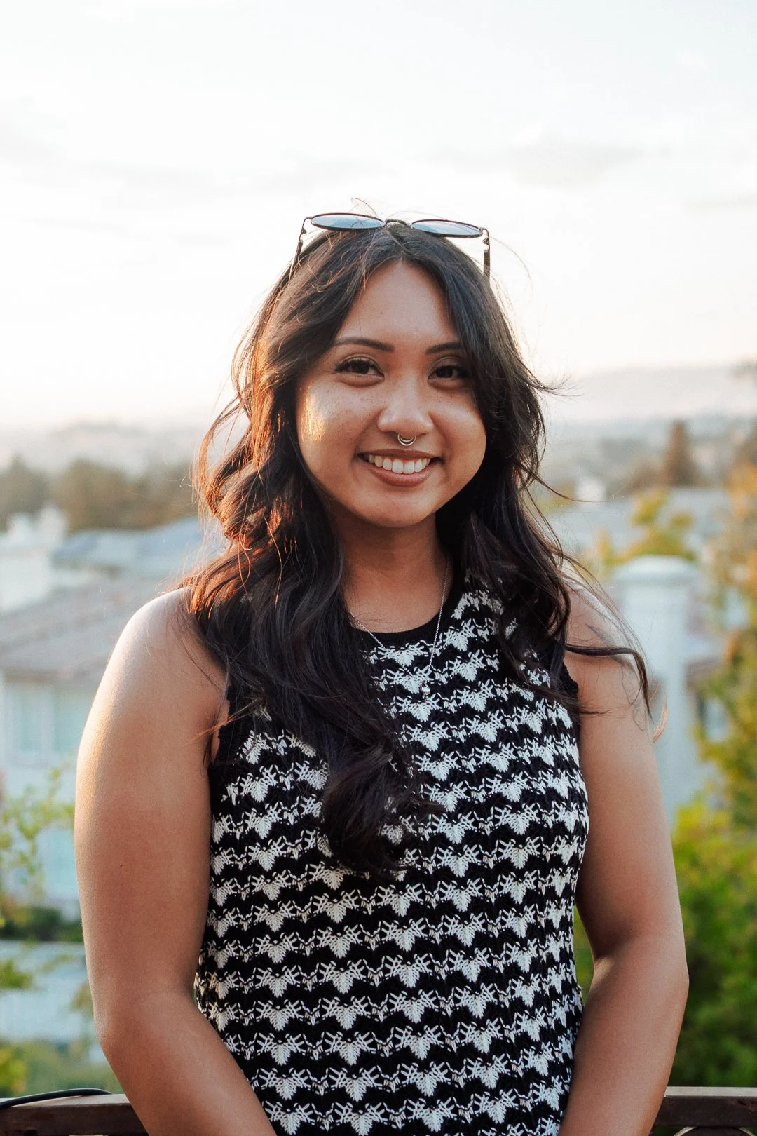 A smiling woman with long dark hair, wearing a black-and-white patterned sleeveless top, sunglasses on her head, and a septum nose ring stands outdoors during sunset, with blurred trees and houses in the background.