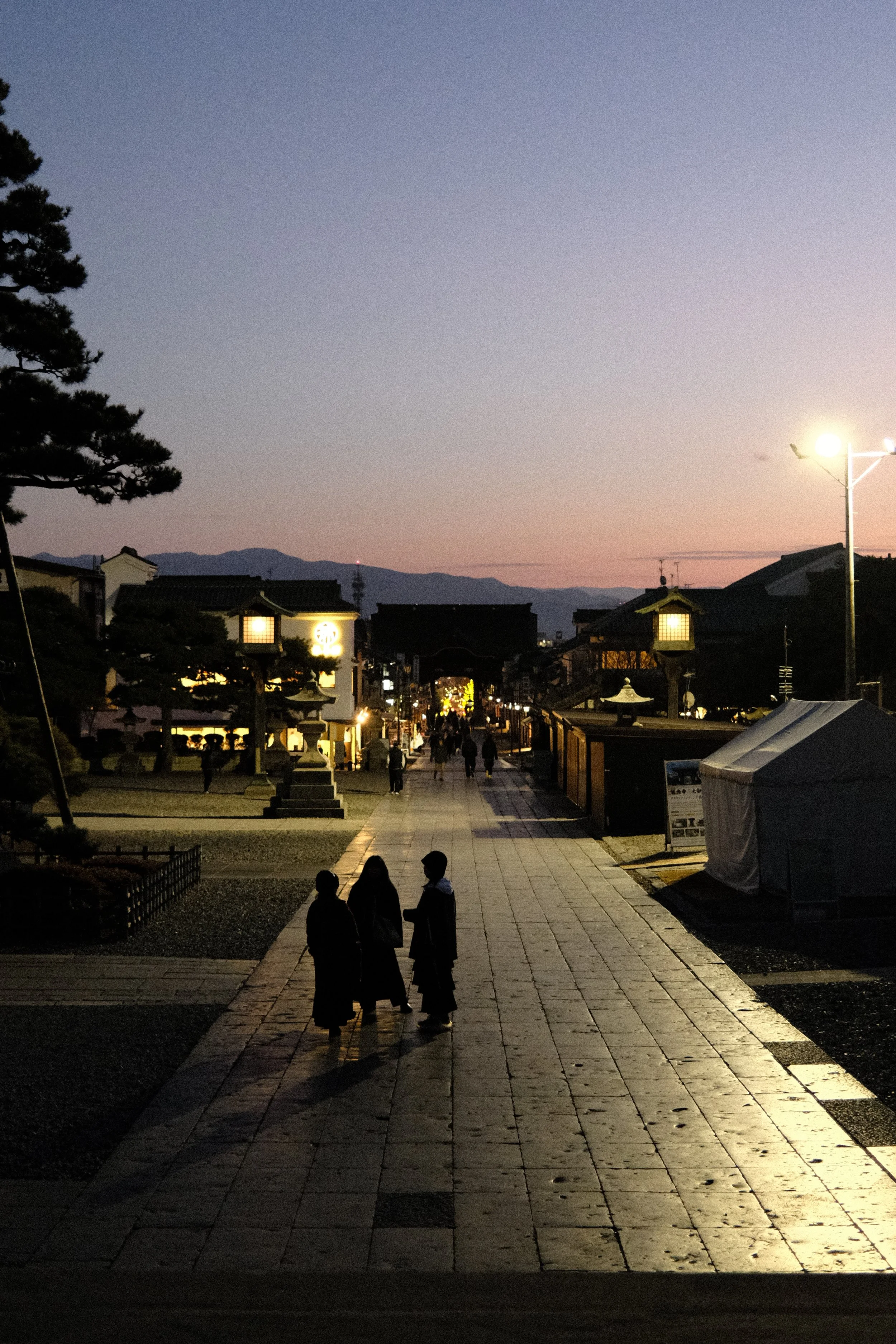 A peaceful street scene at dusk with people walking and traditional lanterns and buildings, with mountains in the background.