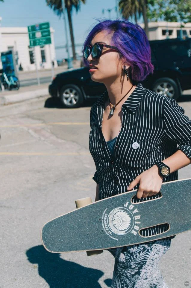 Young woman with purple hair wearing sunglasses, a striped shirt, and patterned pants, holding a skateboard, standing in a parking lot with cars, palm trees, and blue sky in the background.