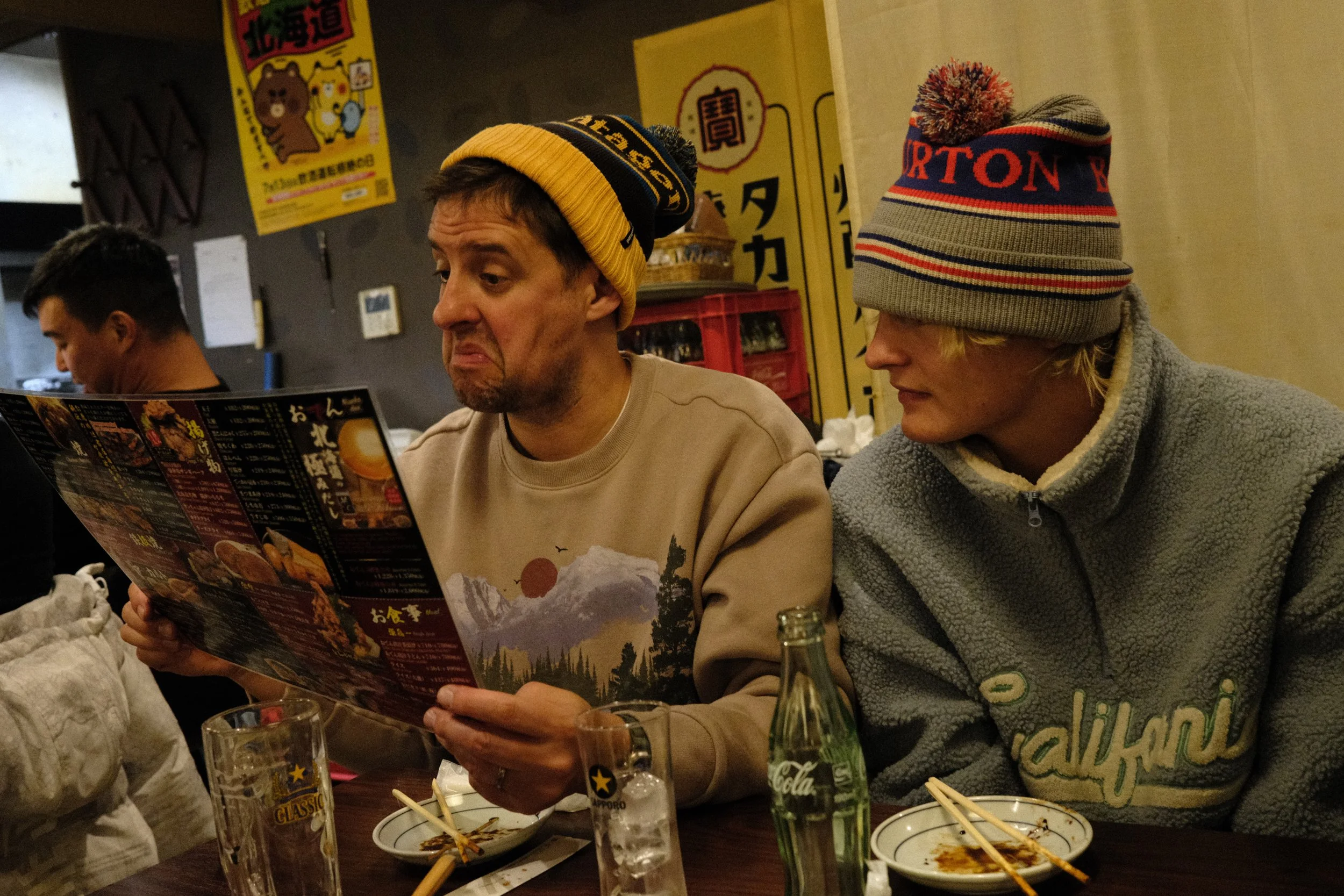 Two men sitting at a restaurant table, looking at a menu. One man is wearing a yellow beanie, and the other is wearing a gray beanie with red and blue stripes.