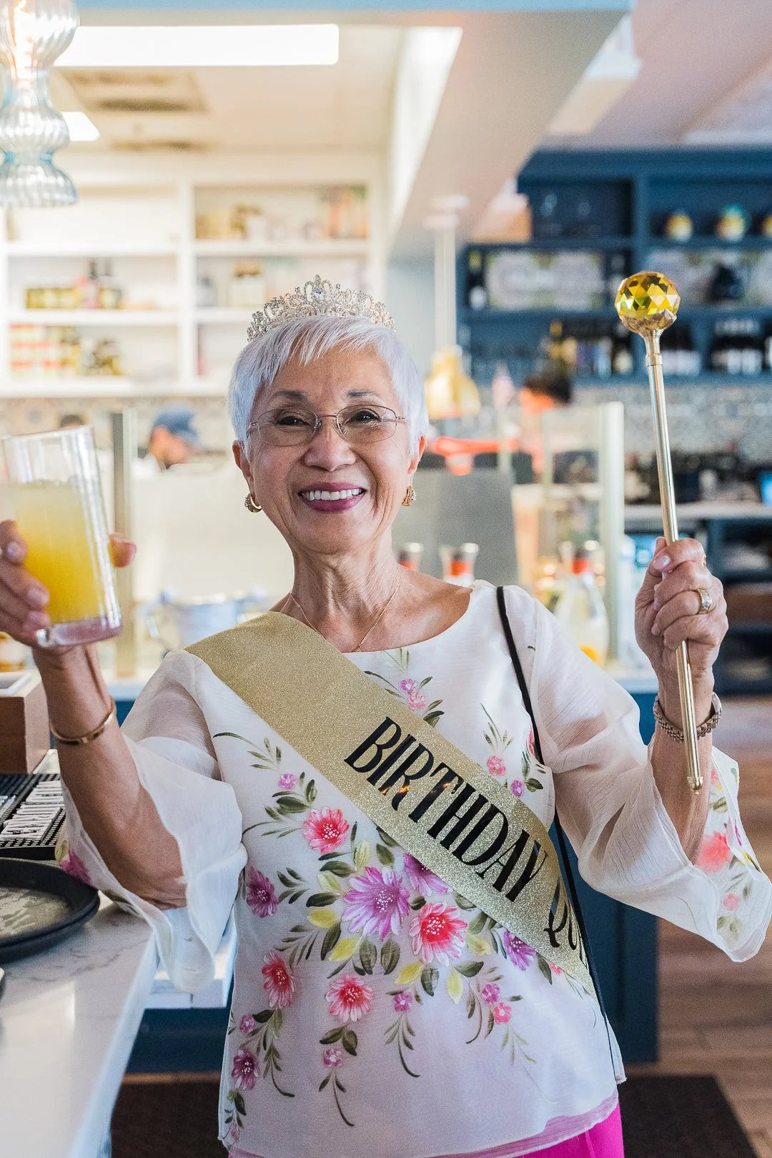 Older woman in a floral blouse with a gold 'Birthday Queen' sash, wearing a crown, and holding a drink and party stick in a festive setting.