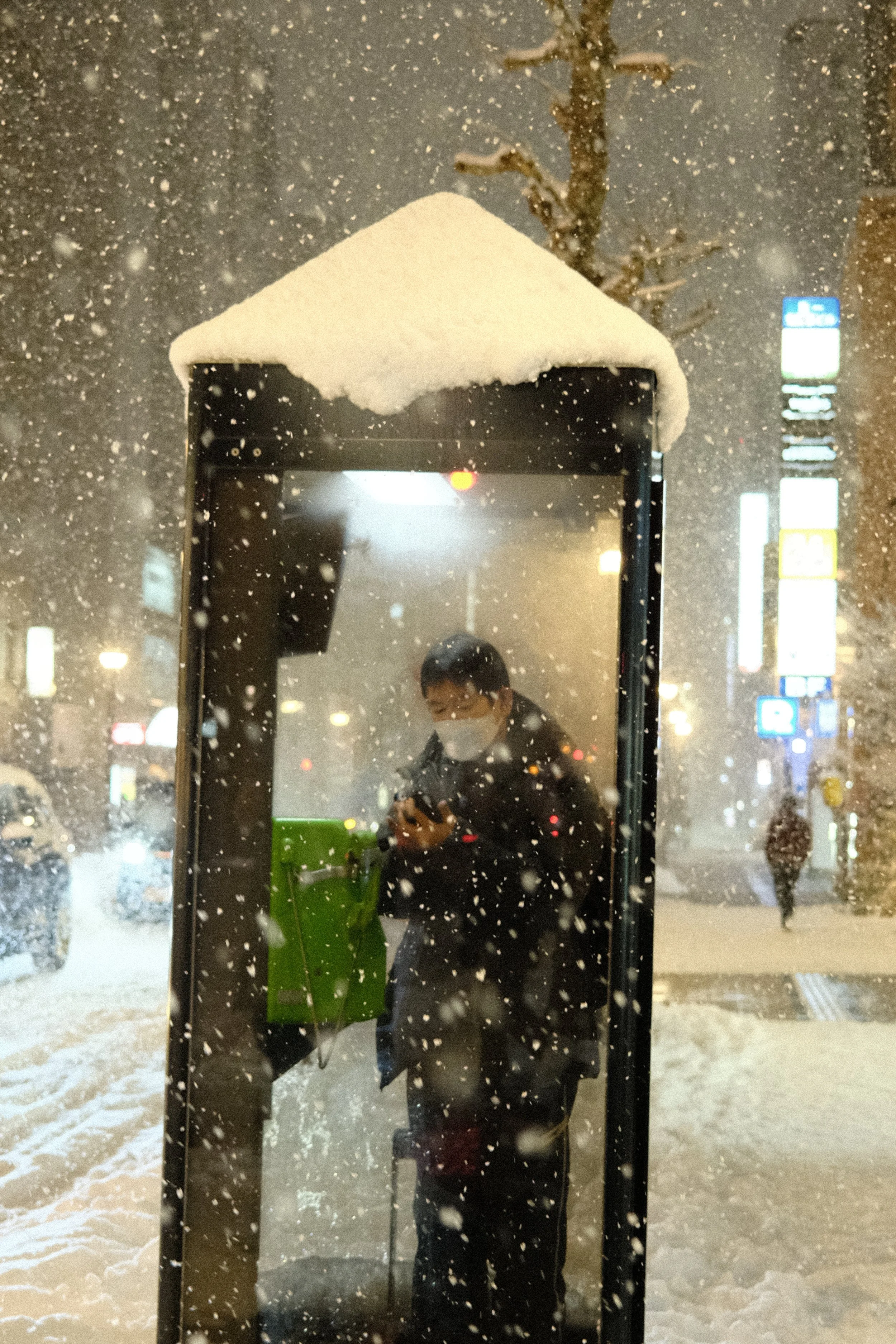 A person wearing a face mask using a public telephone booth in a snowy night in an urban area.