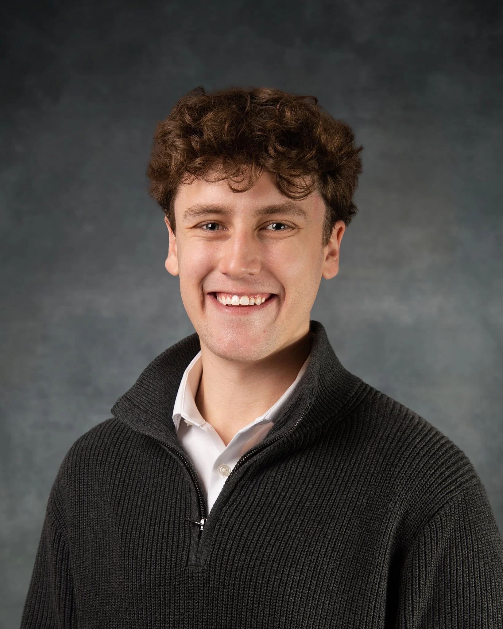 Portrait of a young man with curly brown hair, smiling, wearing a black zip-up sweater over a white collared shirt, against a gray textured background.