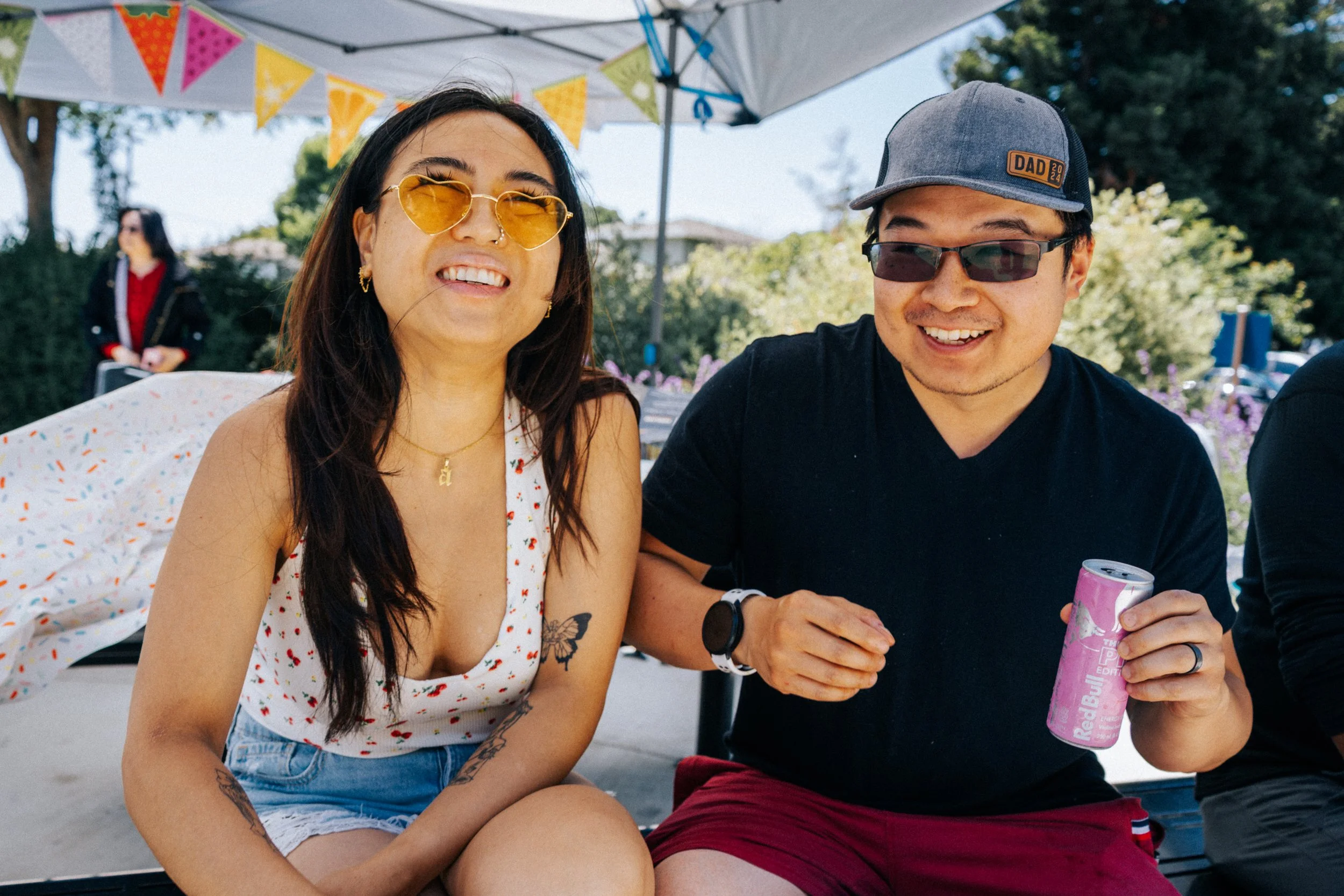 Two friends enjoying outdoor summer event, sitting under a tent with colorful bunting, smiling, one holding a pink can, and the other wearing sunglasses and a cap.