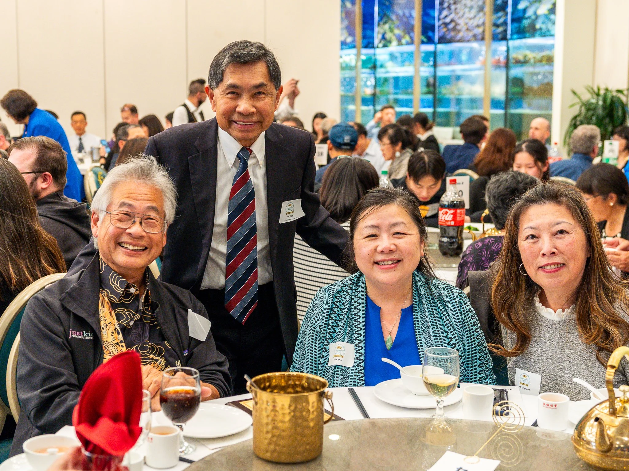 A group of four people sitting at a round table during a formal event, with other attendees in the background. They are smiling and appear to be enjoying themselves. There are drinks, a red napkin, and some tableware on the table.