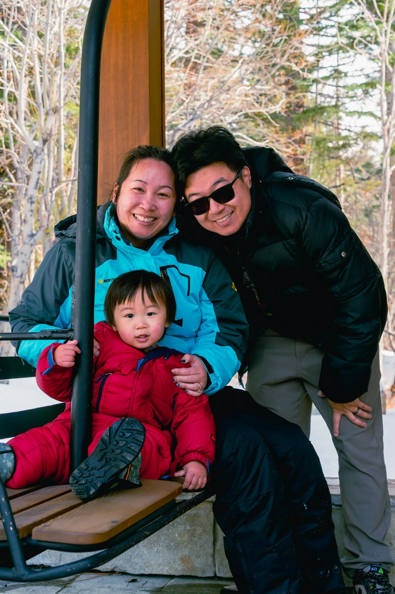 A family of three posing outdoors, with trees in the background. The woman is wearing a blue jacket, the man is wearing sunglasses and black jacket, and the young girl in a red jacket is sitting on a wooden bench.
