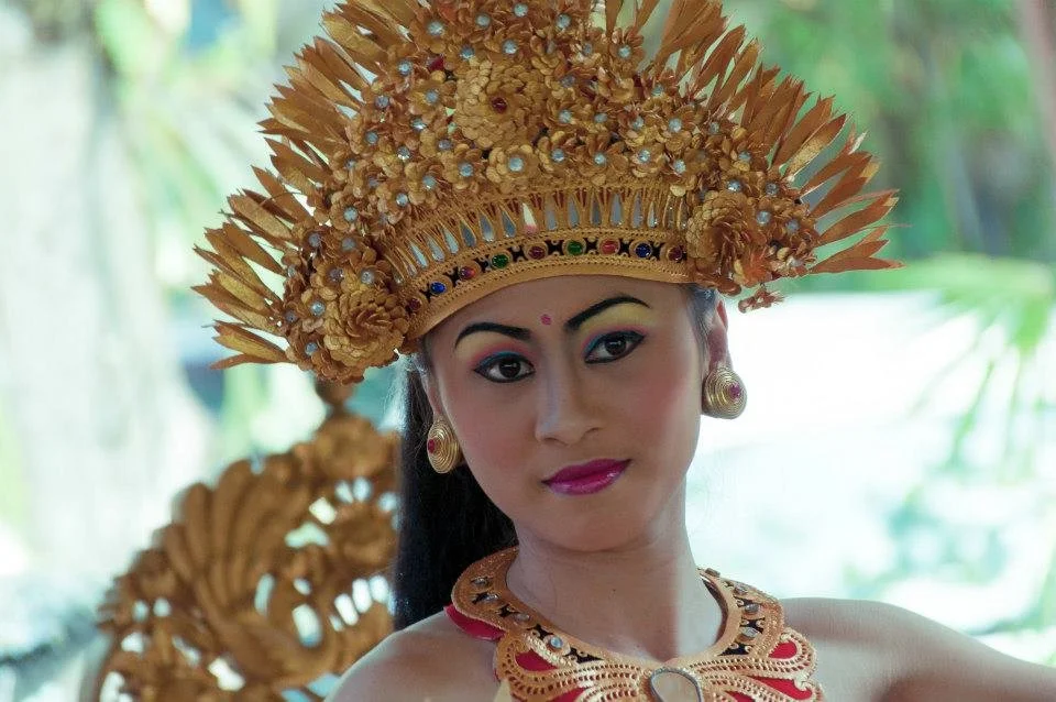 Woman dressed in traditional Balinese attire, wearing an elaborate gold headdress with intricate designs and a necklace with detailed goldwork, against a natural outdoor background.