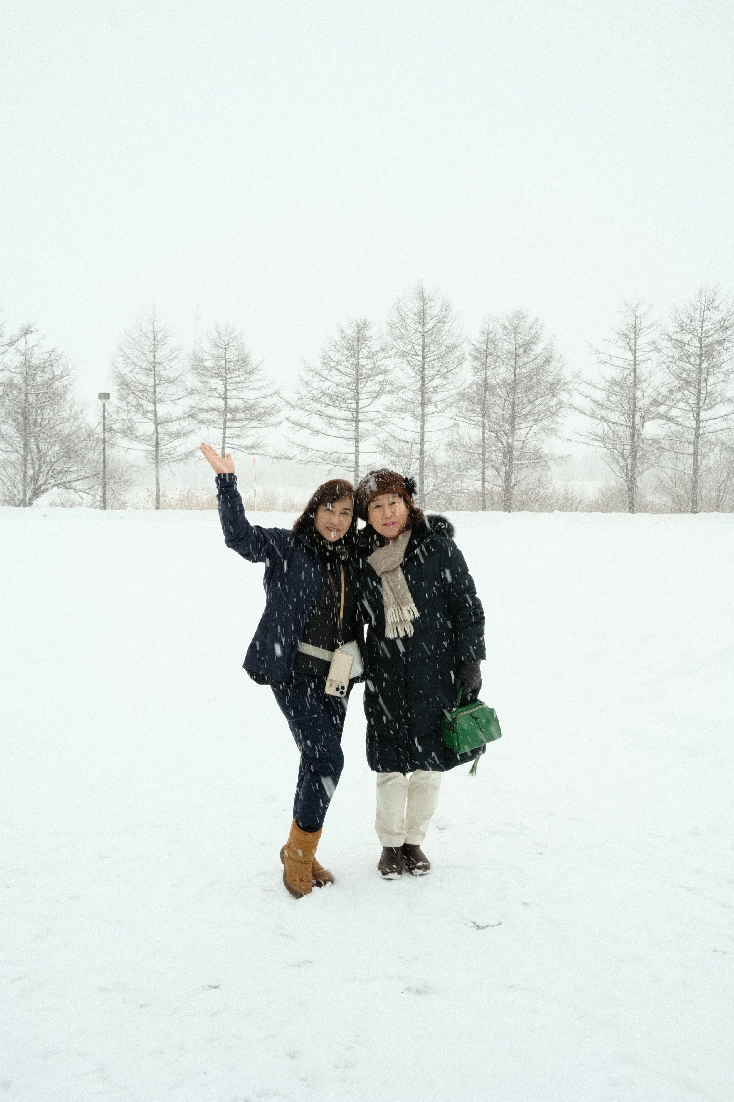 Two women standing in the snow on a winter day, surrounded by snow-covered trees, with one woman raising her right hand in the air.