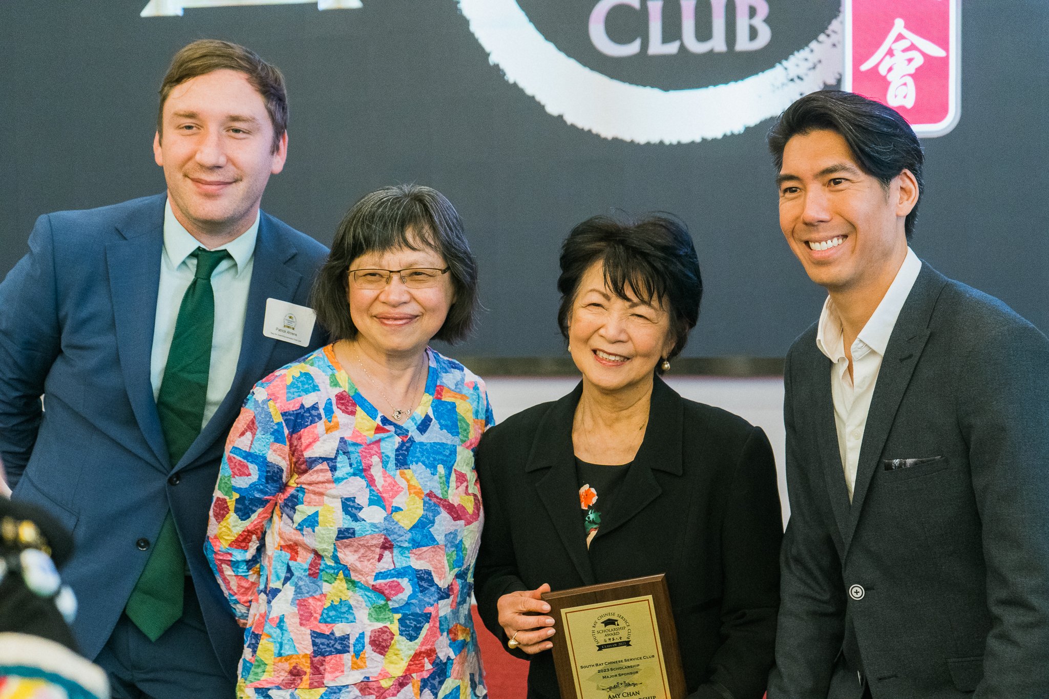 Four people posing and smiling for a photo at an award ceremony, with a woman in the center holding a plaque. The background features a black and pink sign that says 'CLUB' and some Japanese characters.