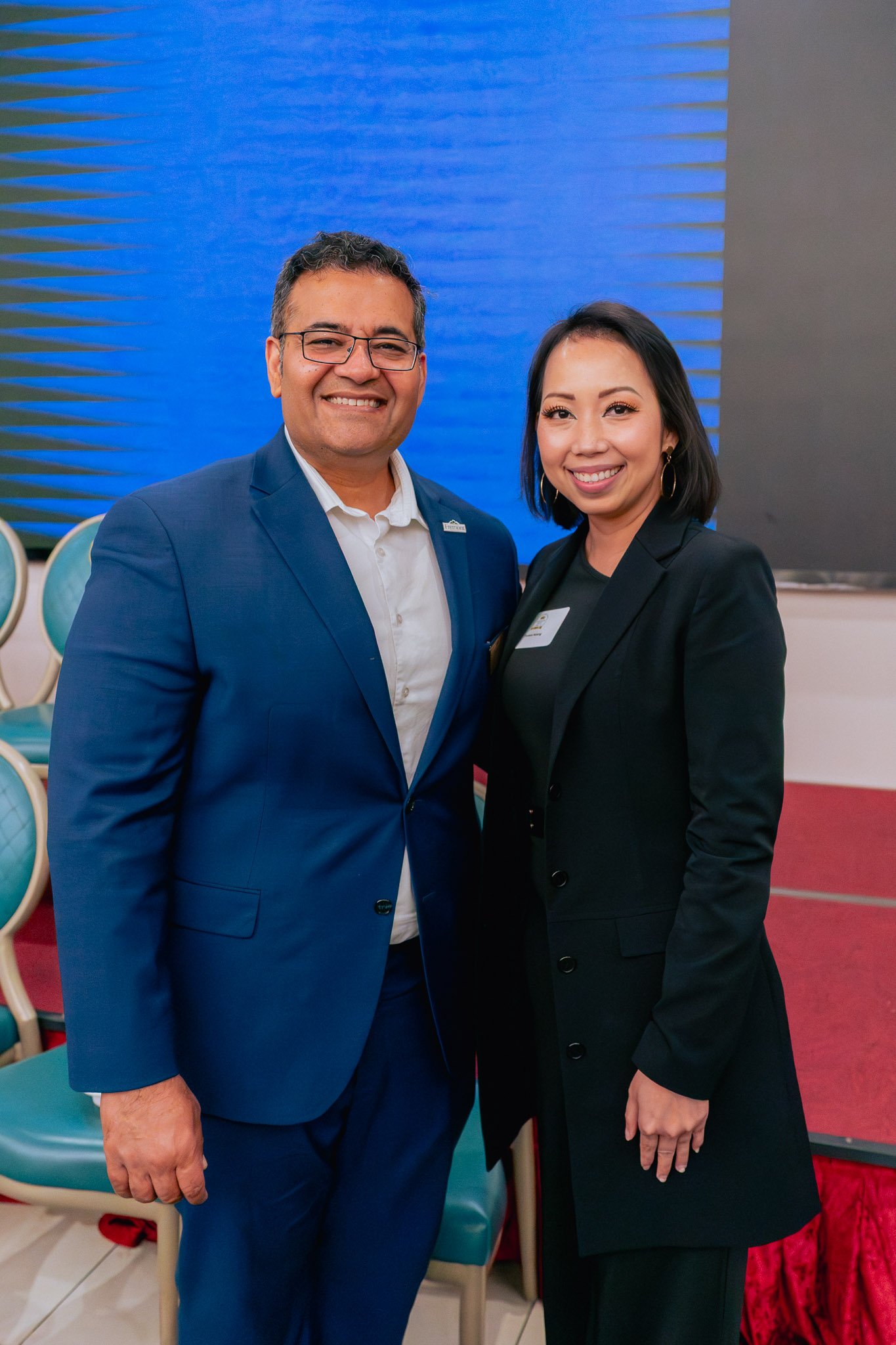 Two smiling professionals, a man in a navy blue suit and a woman in a black blazer, posing indoors with stacked chairs in the background.