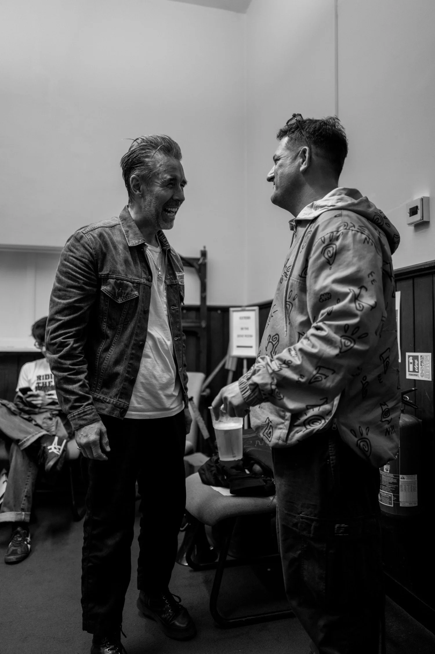 Two men are standing and laughing at each other in an empty room. One is wearing a denim jacket and the other a hoodie with patterns.