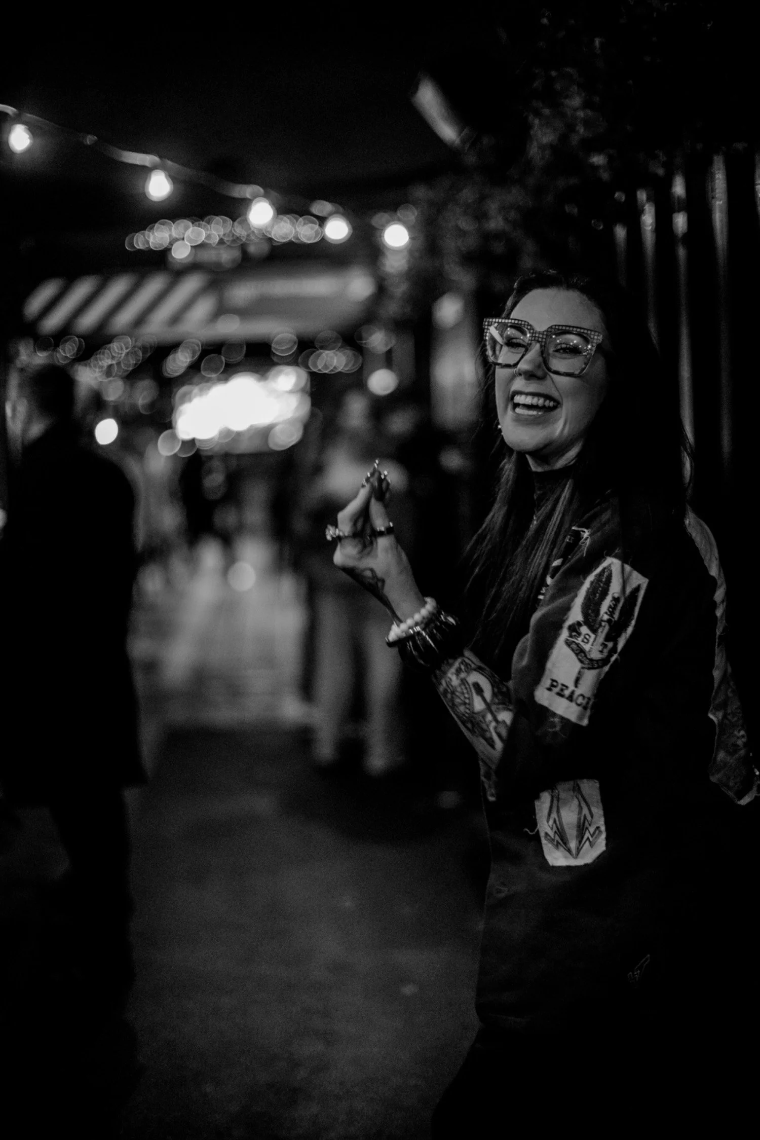 A woman with long dark hair, wearing large glasses with rhinestones, smiling and holding a small object in her hand, standing on a dimly lit street at night with string lights overhead and blurred people in the background.