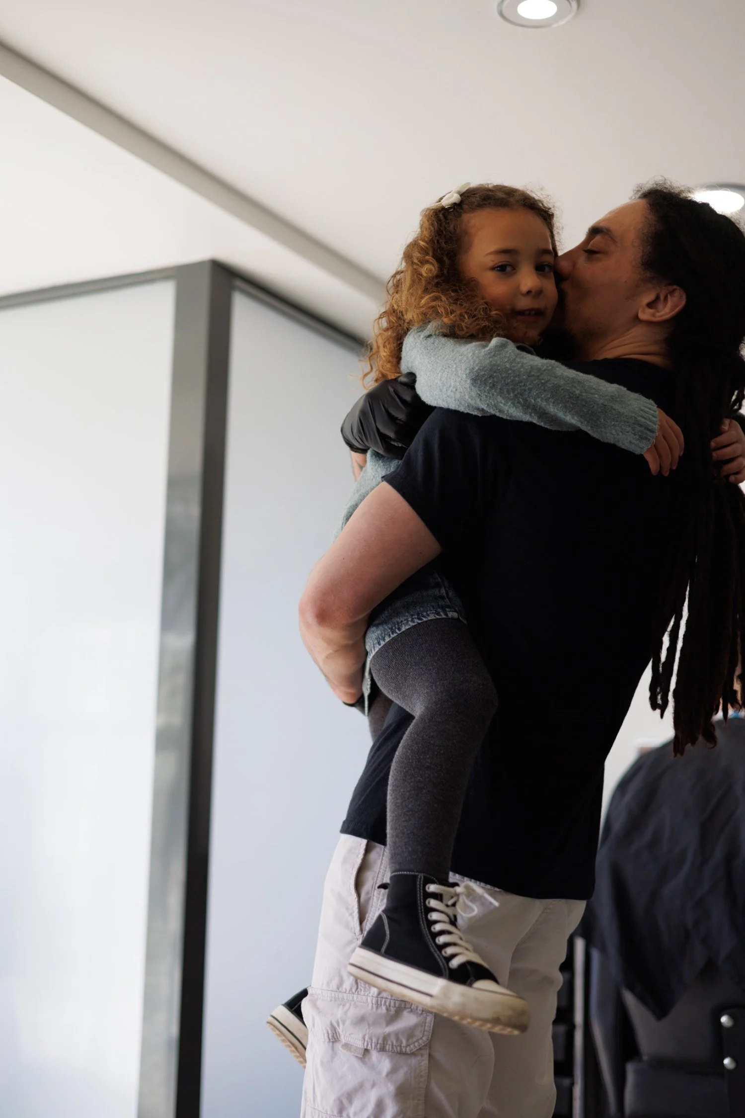 A candid moment of a man holding a young girl on his shoulder, kissing her cheek indoors with white walls and ceiling lights.