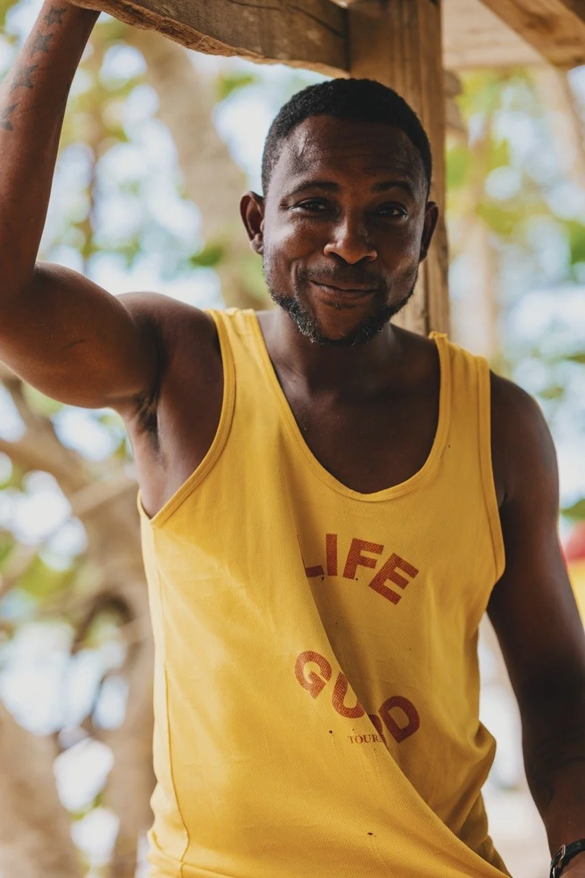 A man wearing a yellow tank top with red text, smiling and leaning against a wooden structure outdoors with blurred trees in the background.