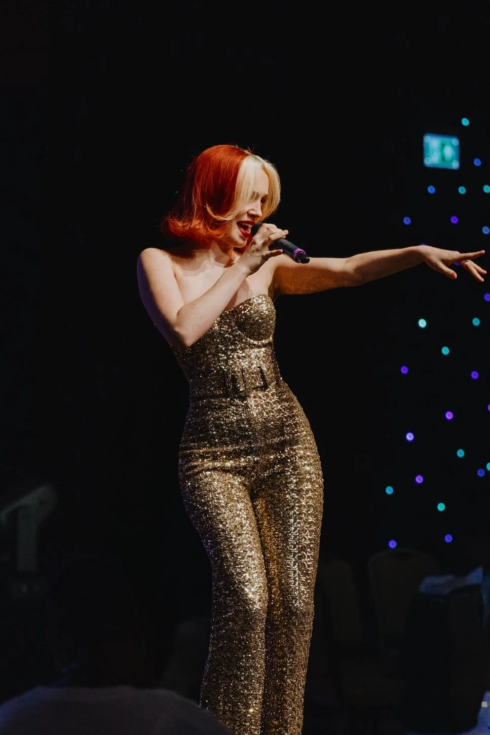 A woman performing on stage, wearing a glittery gold jumpsuit, holding a microphone, with her arm extended, against a dark background with colorful lights.