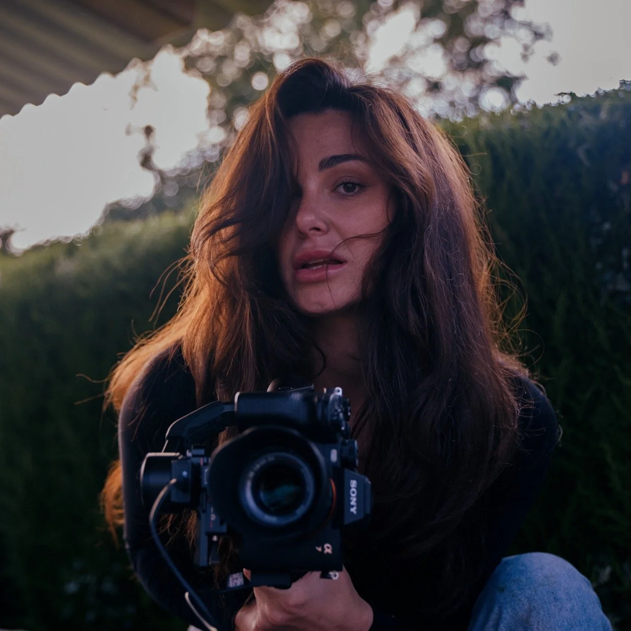 A woman with long dark hair holding a camera, sitting outdoors near greenery with a blurred background.