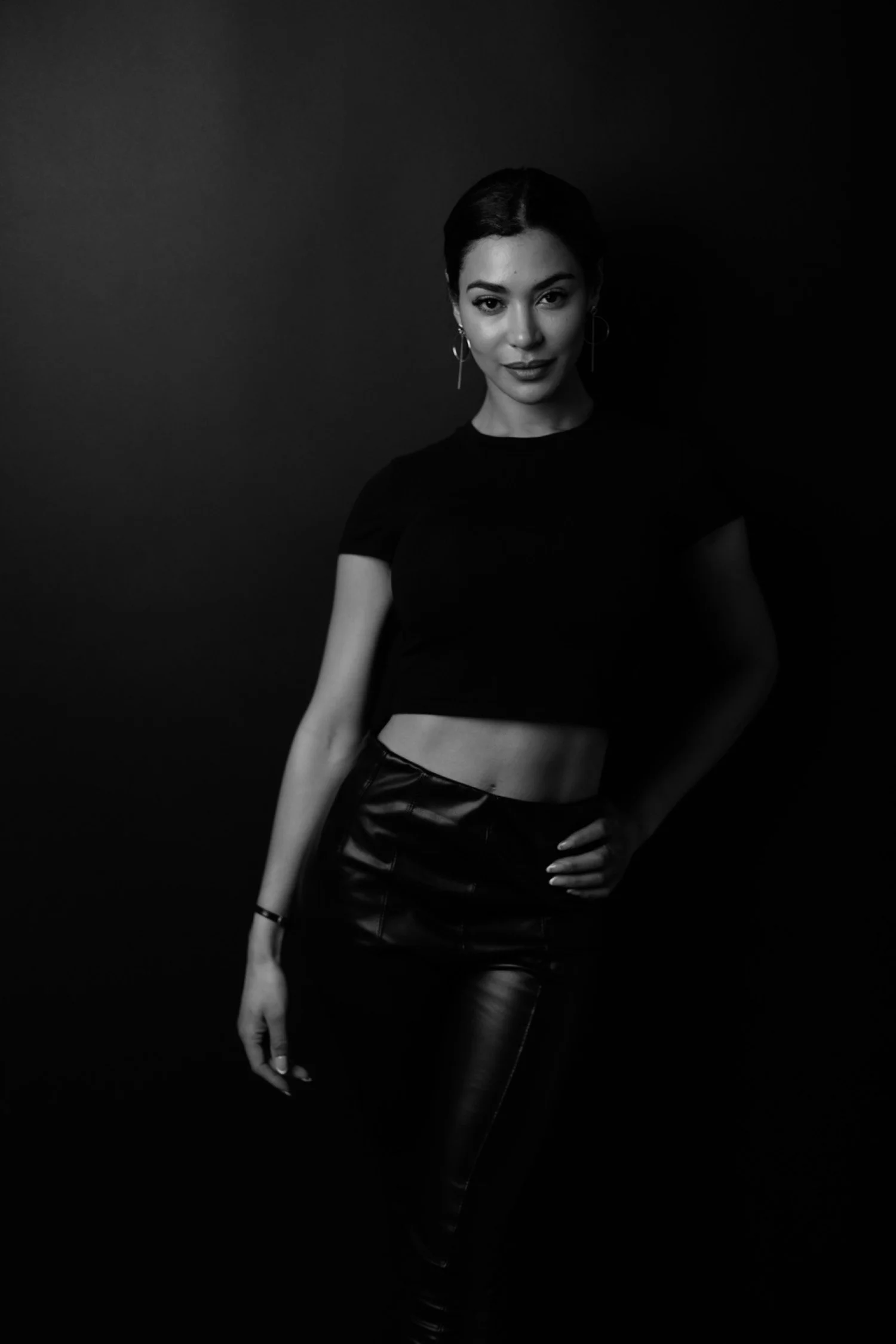 A woman with short dark hair and hoop earrings posing against a dark background, wearing a black crop top and high-waisted leather pants.