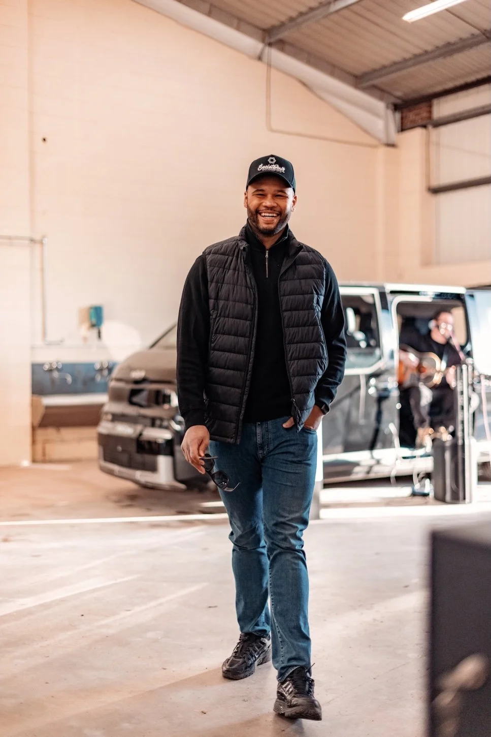 A smiling man is walking in an indoor space, holding sunglasses in his right hand. He is dressed in a black jacket, jeans, and a cap. In the background, a person is sitting with a guitar.