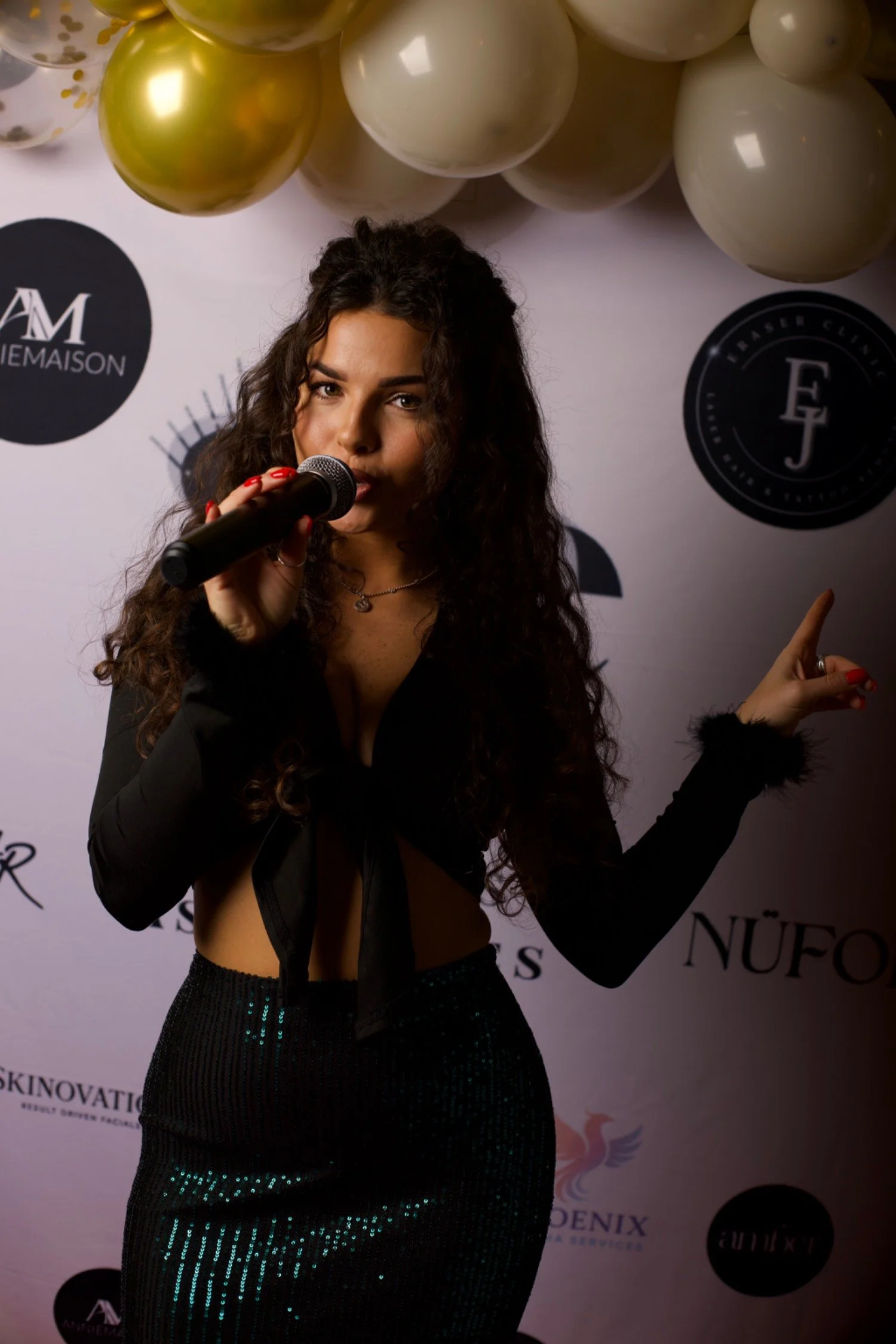 A woman with long curly hair holding a microphone and pointing to the side with her other hand, standing in front of a backdrop decorated with gold and white balloons and logos.