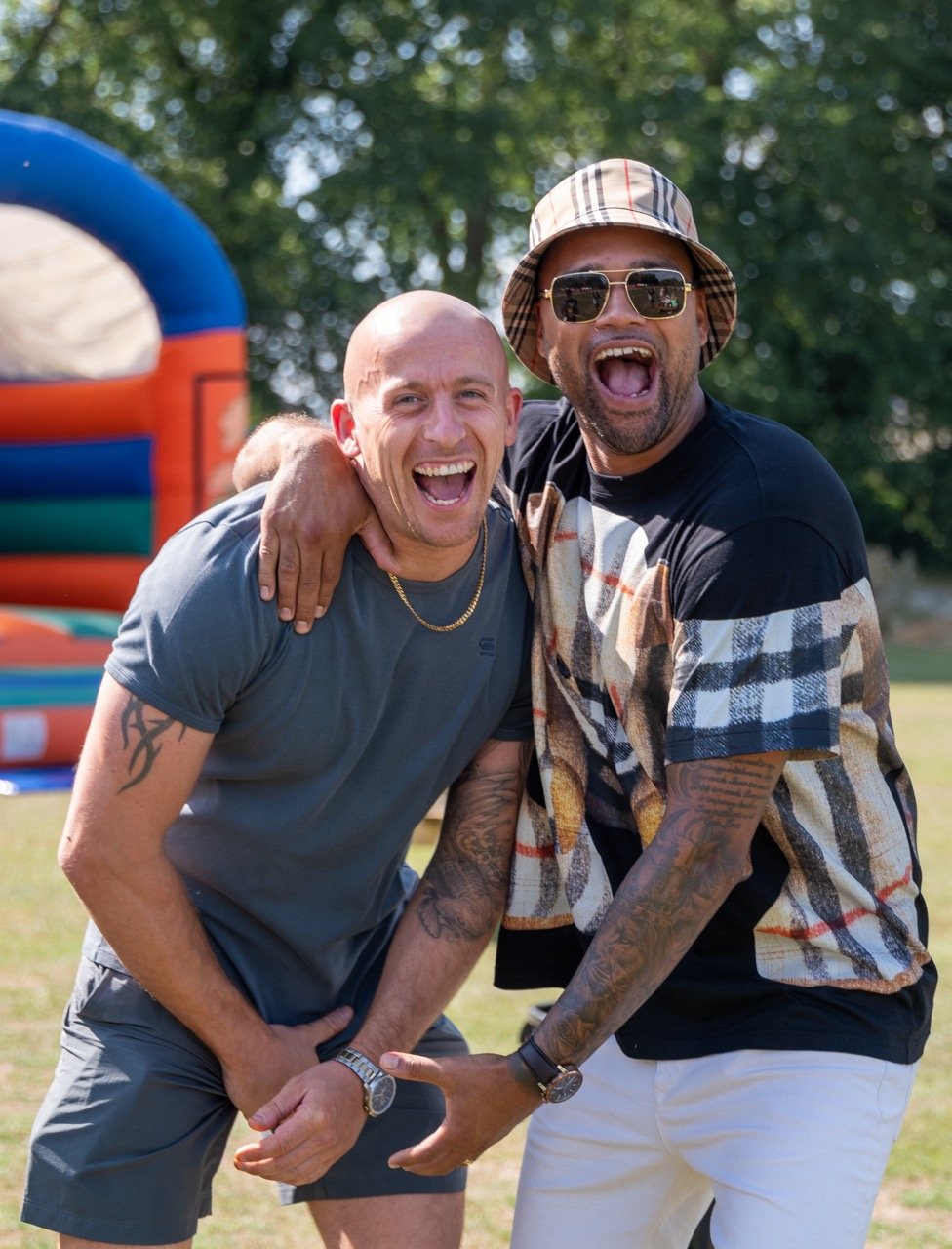 Two men smiling and laughing outdoors, one with a shaved head and tattoos, wearing a gray t-shirt, and the other with a beard, sunglasses, and a bucket hat, wearing a patterned shirt. They are standing close together with one man's arm around the oth