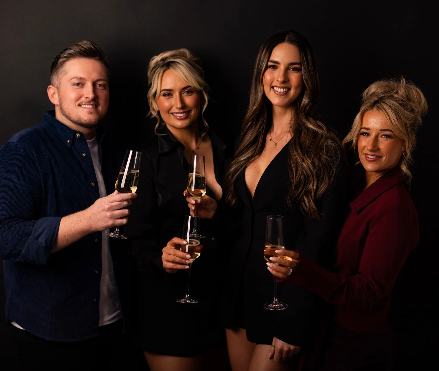 Four people dressed in semi-formal attire, holding glasses of champagne, standing close together against a black background, smiling at the camera.