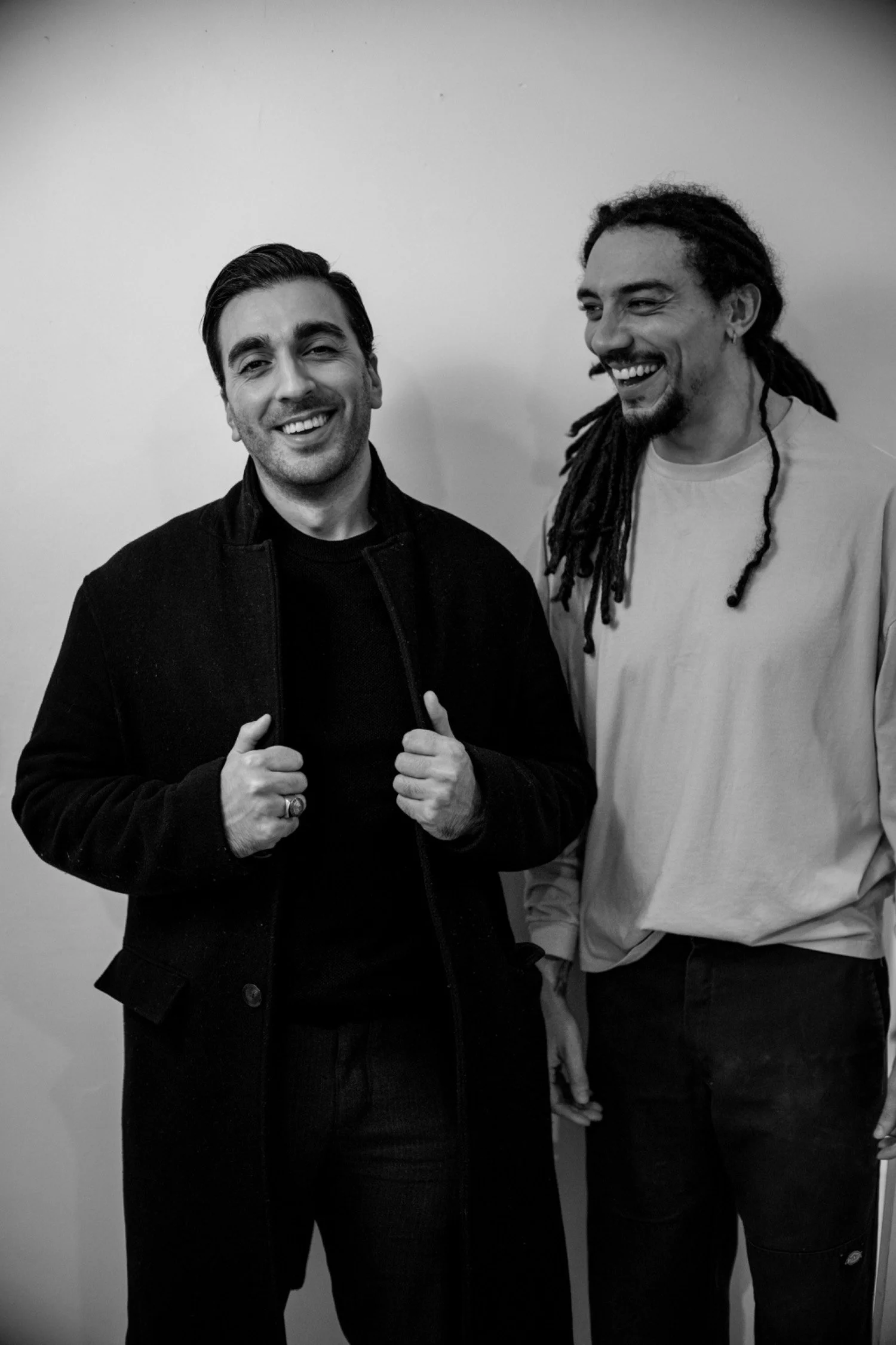 Two men smiling and standing side by side in front of a plain wall. One man has dark hair, is wearing a dark jacket, and holding the collar with both hands. The other man has dreadlocks, a beard, and is wearing a light-colored long-sleeve shirt. Both