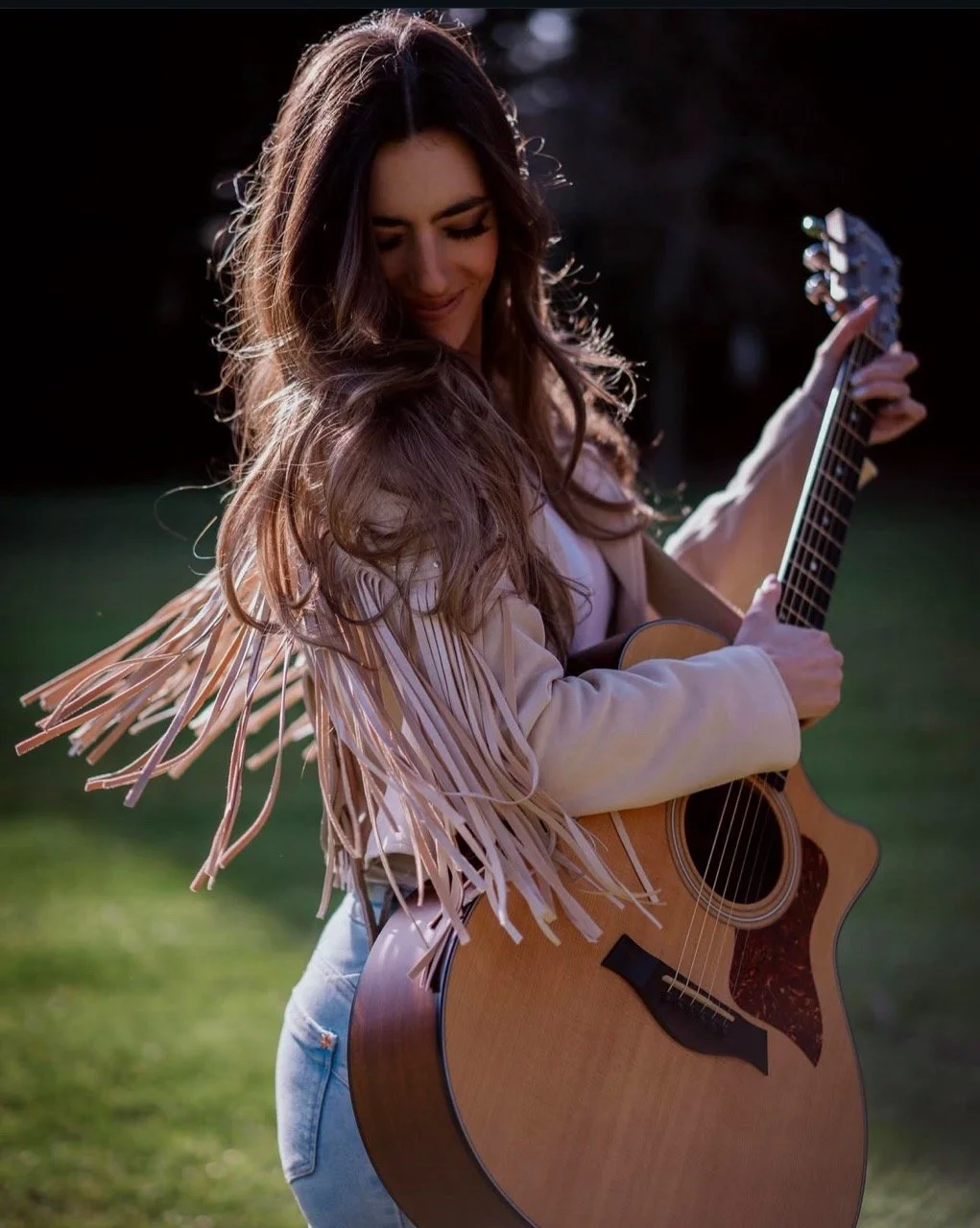 Woman playing an acoustic guitar outdoors, smiling, with long wavy hair, wearing a fringed jacket.