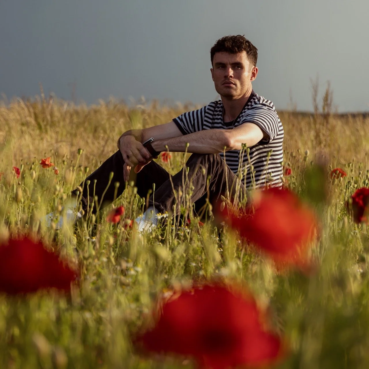 A young man with short curly hair sitting in a field of red poppies during sunset, wearing a striped t-shirt and black pants, looking at the camera.