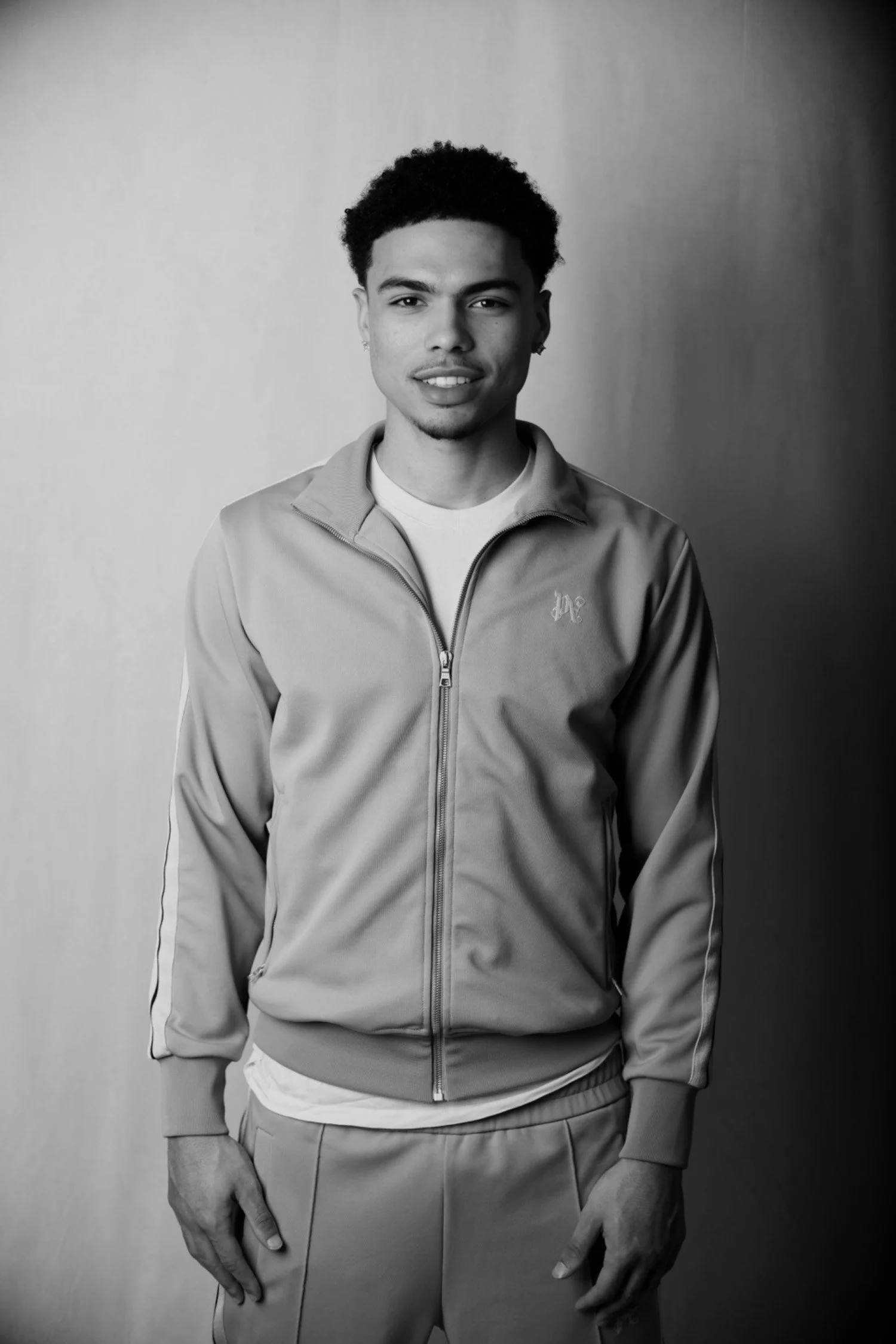 Black and white photo of a young man with short curly hair, wearing a zip-up jacket and pants, standing against a plain background.