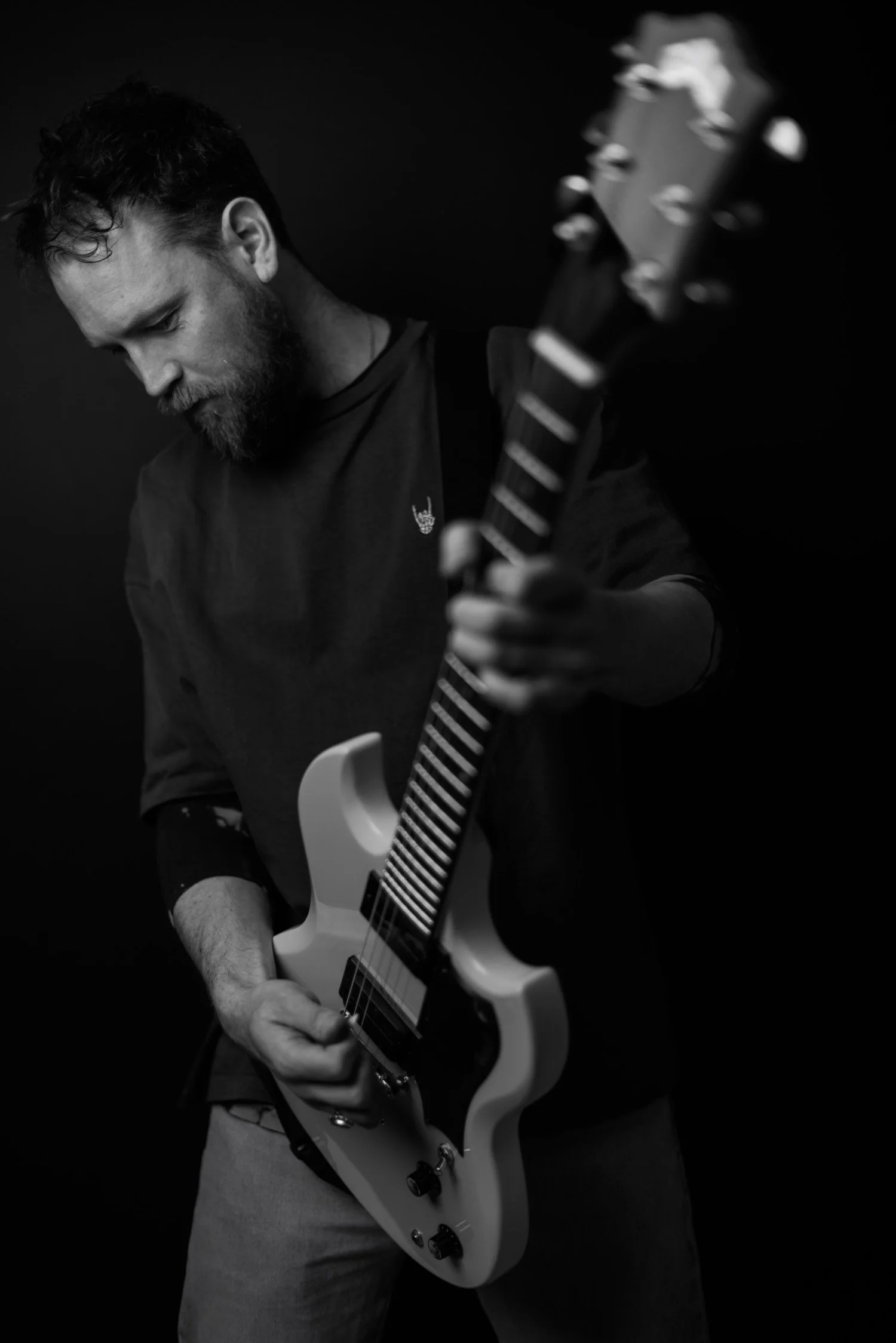 Black and white image of a man with a beard and short, curly hair, playing an electric guitar. He is wearing a dark sweatshirt and light-coloured trousers, with a small pin on his shirt. The background is plain and dark.