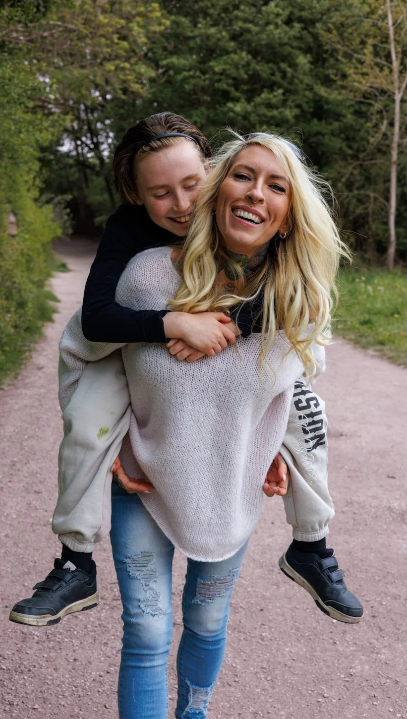 Candid photo of a mum and boy, both are smiling as the mum gives the boy a piggyback ride on a dirt path surrounded by green trees.
