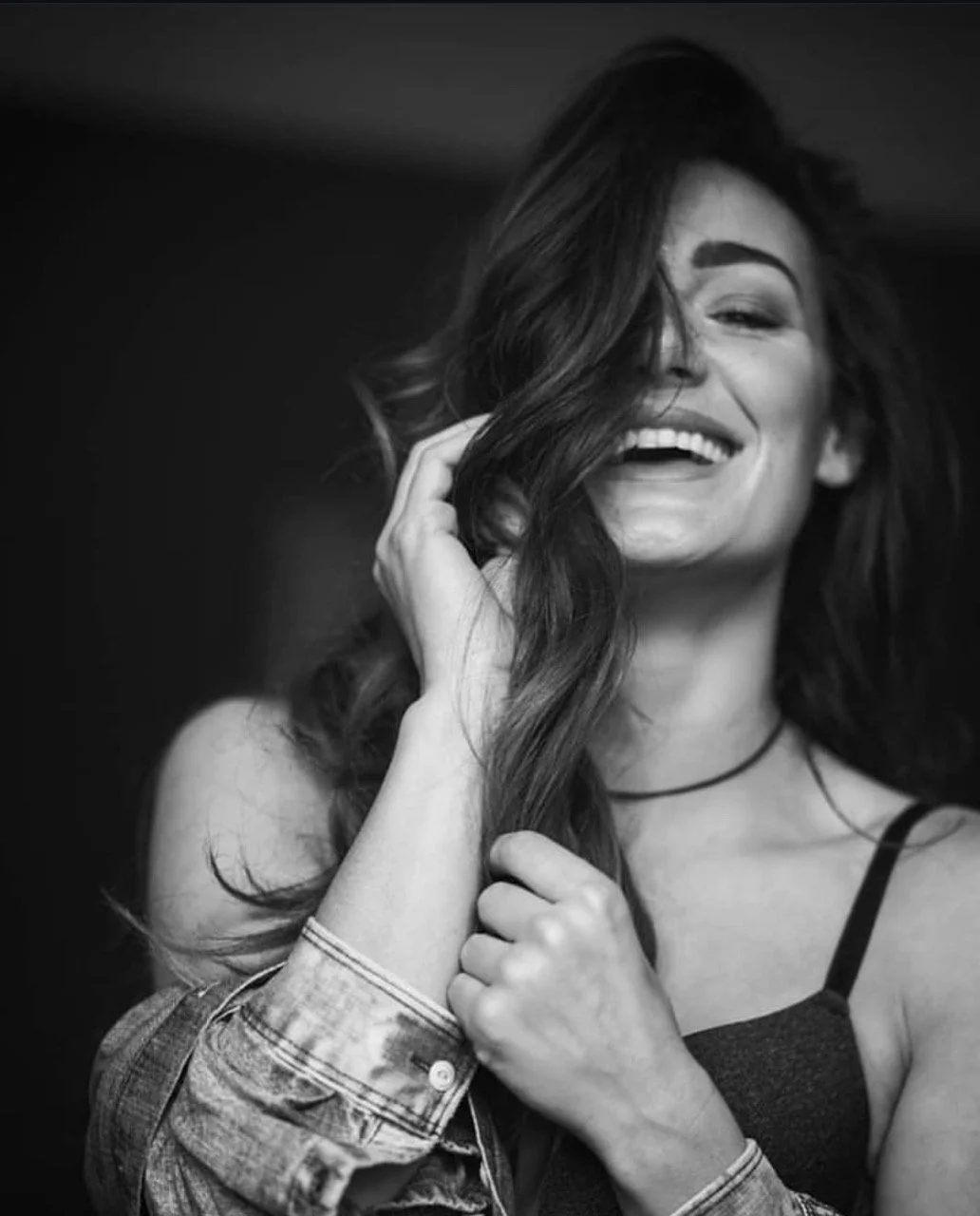 A smiling woman with long wavy hair, dressed in a sleeveless top, holding her hair with one hand and touching her face with the other, in a candid black-and-white photograph.
