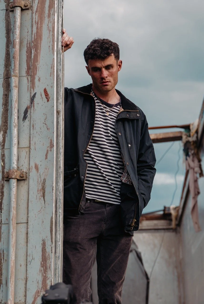 A young man with short curly hair and a serious expression stands outside in front of a weathered, rusted structure. He wears a dark jacket over a black and white striped shirt, with a necklace visible around his neck. The background features a cloud