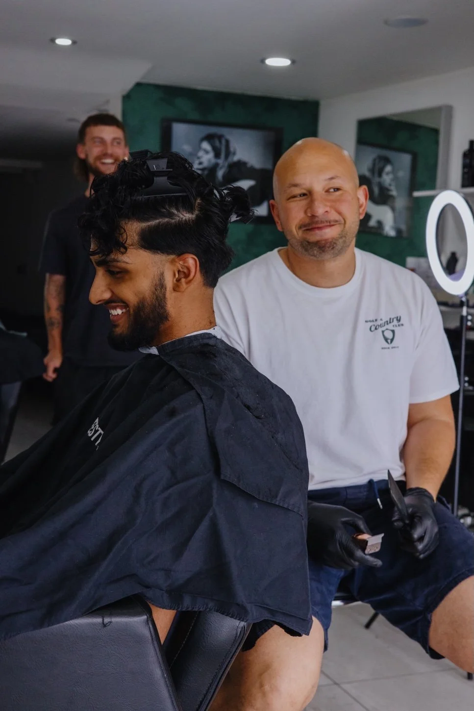 A man with a beard getting a haircut at a salon, smiling. The barber, a bald man wearing a white T-shirt and black gloves, is standing behind him. Another man with long hair and a beard is visible in the background, also smiling.