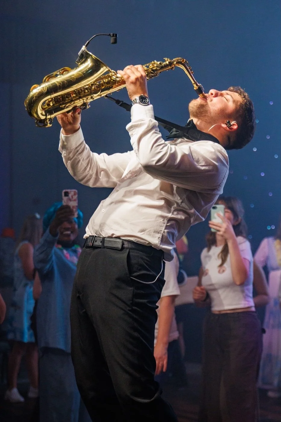 A man wearing a white shirt and black pants plays a gold saxophone at a party, surrounded by people taking photos.