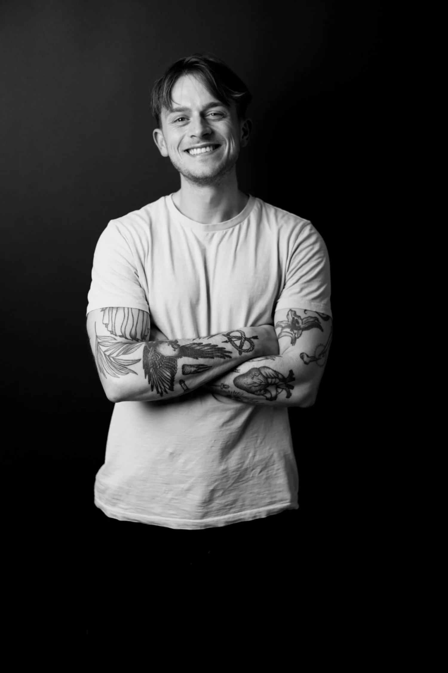 A smiling young man with tattoos on his arms, wearing a light-colored t-shirt, standing with arms crossed against a dark background.