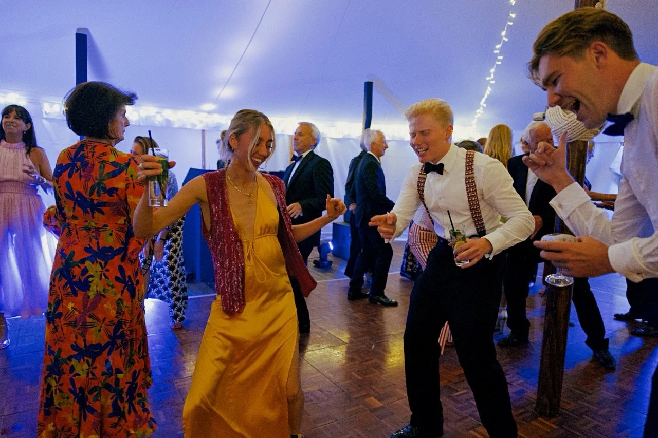 People dancing and laughing at a party under a tent with string lights, dressed in formal and colourful attire.