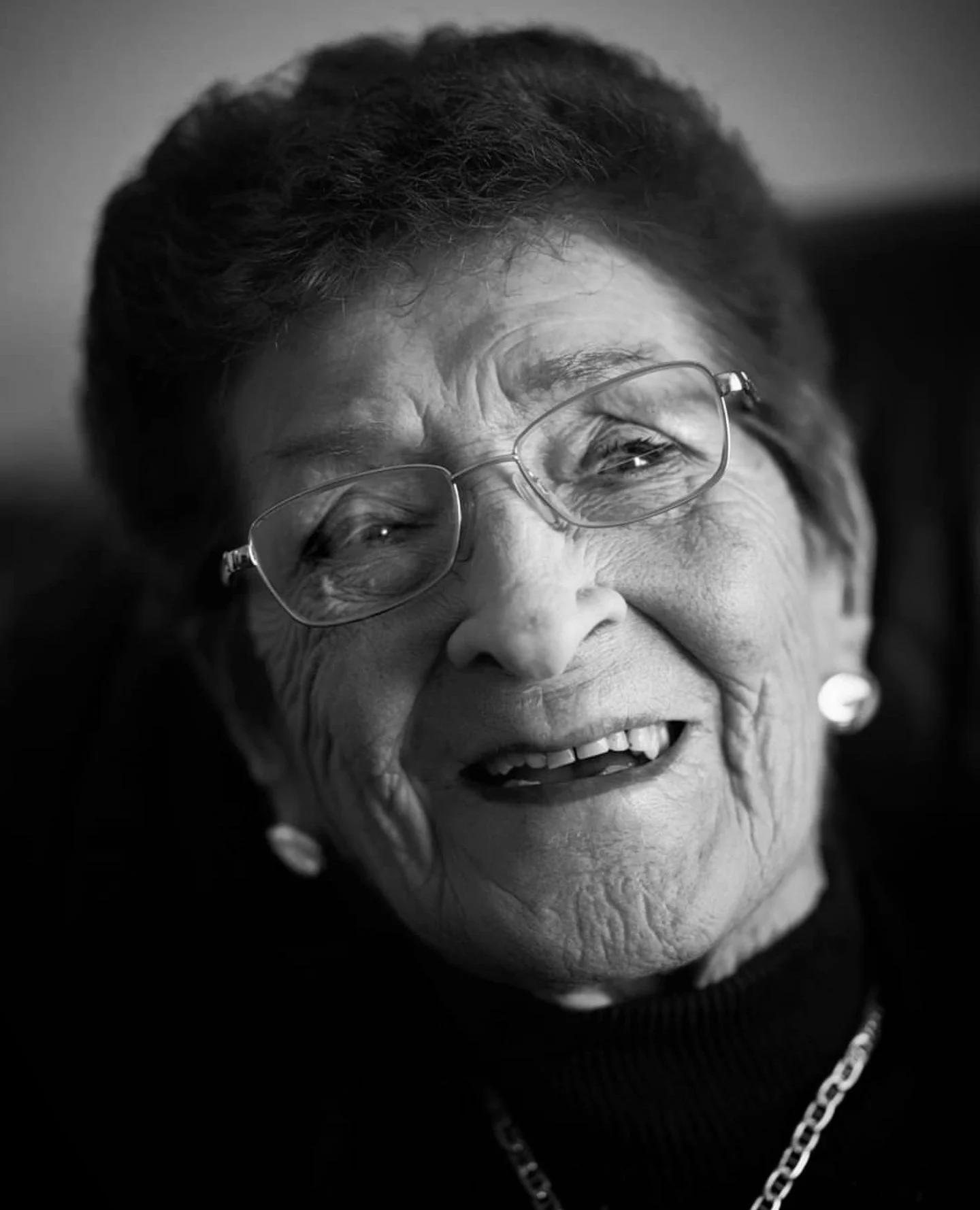 Close-up black and white portrait of an elderly woman smiling, wearing glasses, earrings, and a chain necklace.
