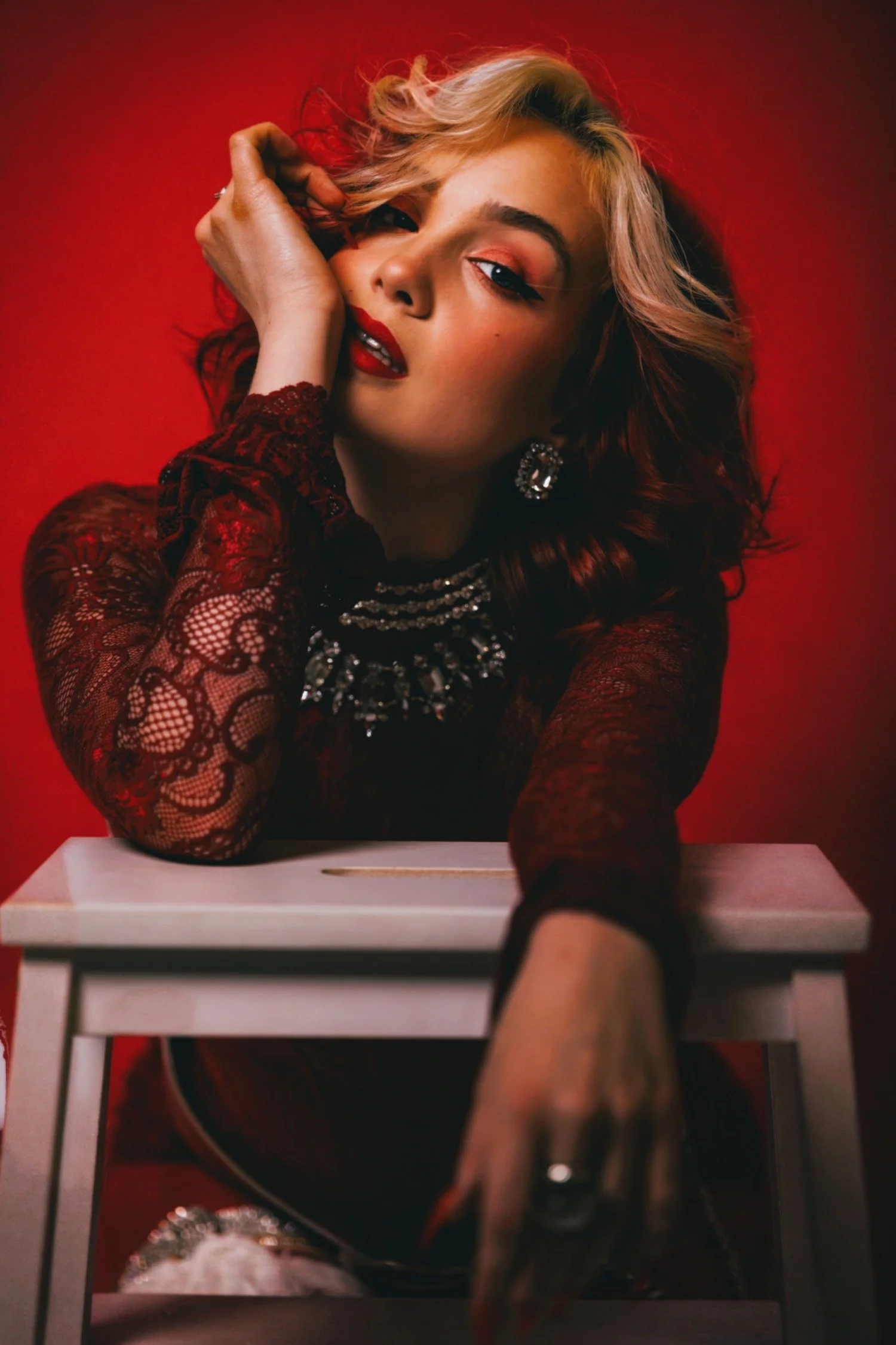 A woman with blonde, wavy hair and red lipstick is sitting in front of a red backdrop. She is wearing a black lace top with pearl jewelry and large earrings. She has one hand near her face and is taking a selfie with a smartphone, which is being held by her outstretched arm.