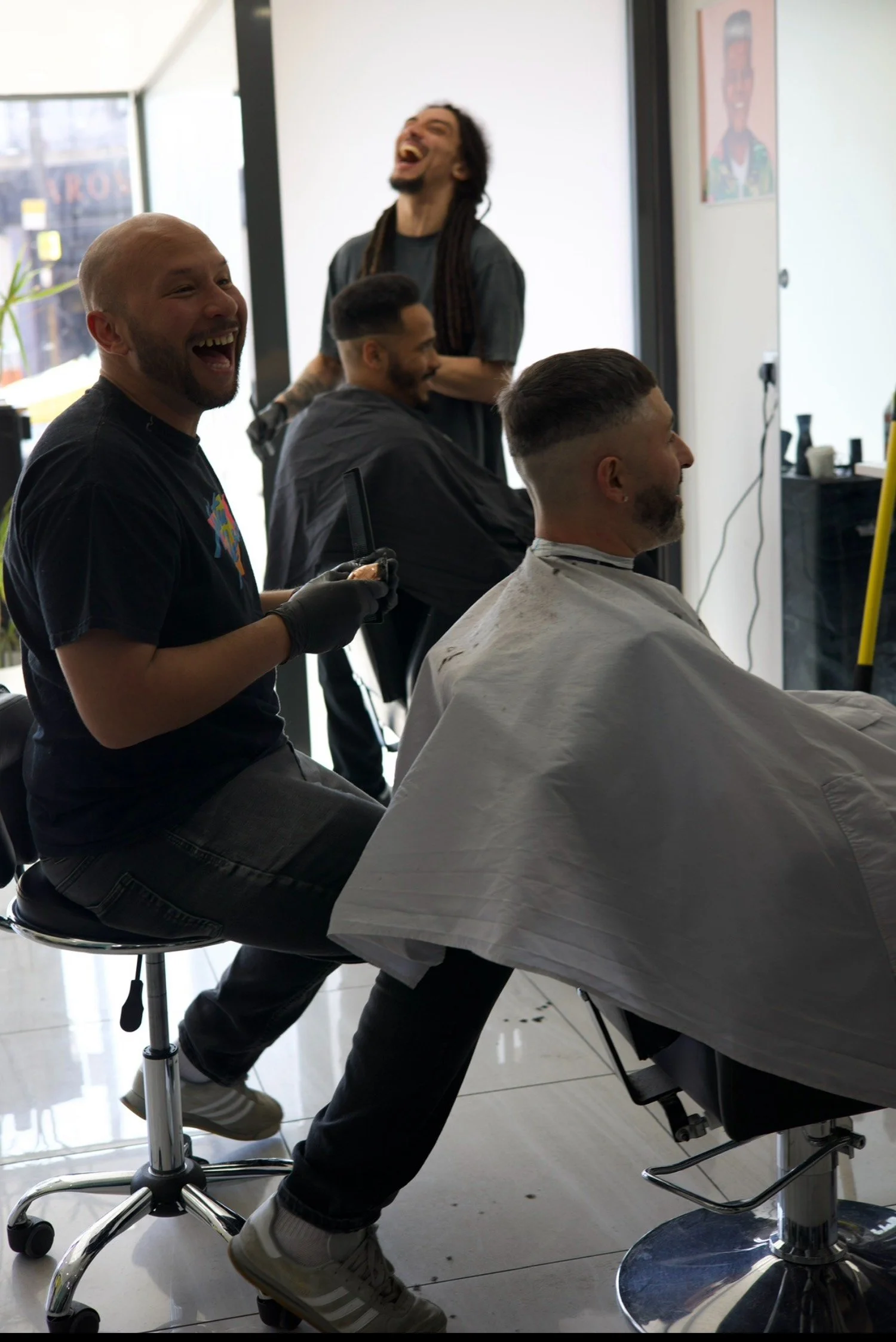 Barber cutting a man's hair while two other men sit in chairs, all laughing in a barber shop.