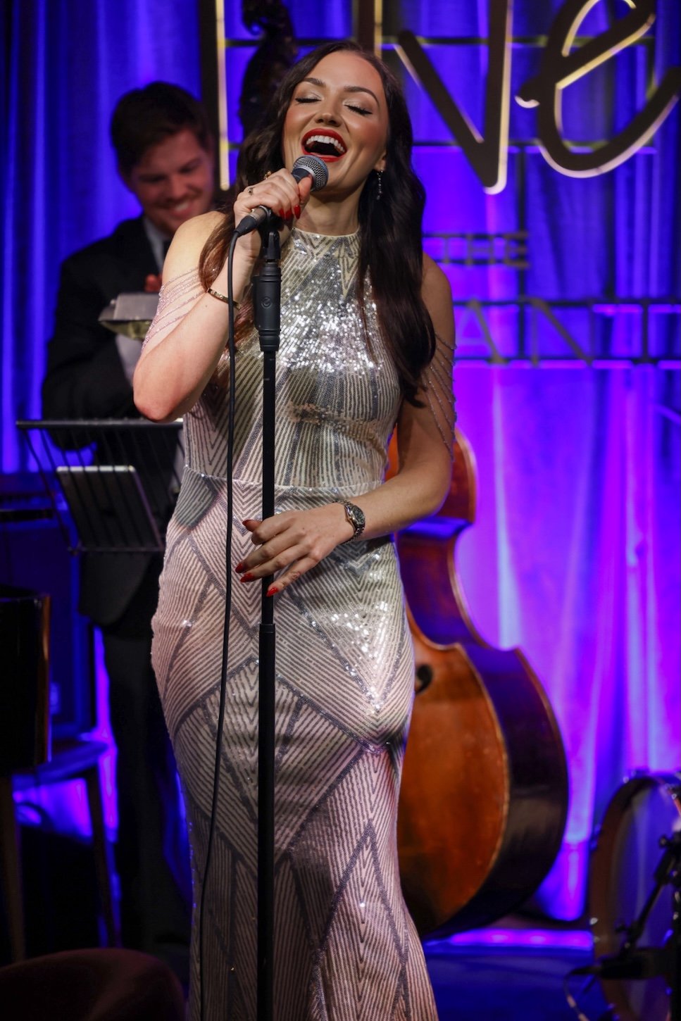 A woman singing into a microphone on stage, wearing a glittery silver gown, with a bass and a musician playing a keyboard in the background, illuminated by purple stage lights.