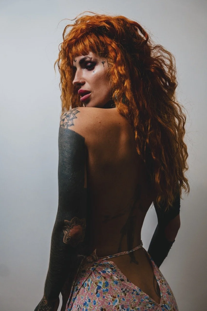 A woman with long, curly red hair looking over her shoulder, with tattoos on her arm and back, wearing a floral skirt and black gloves.