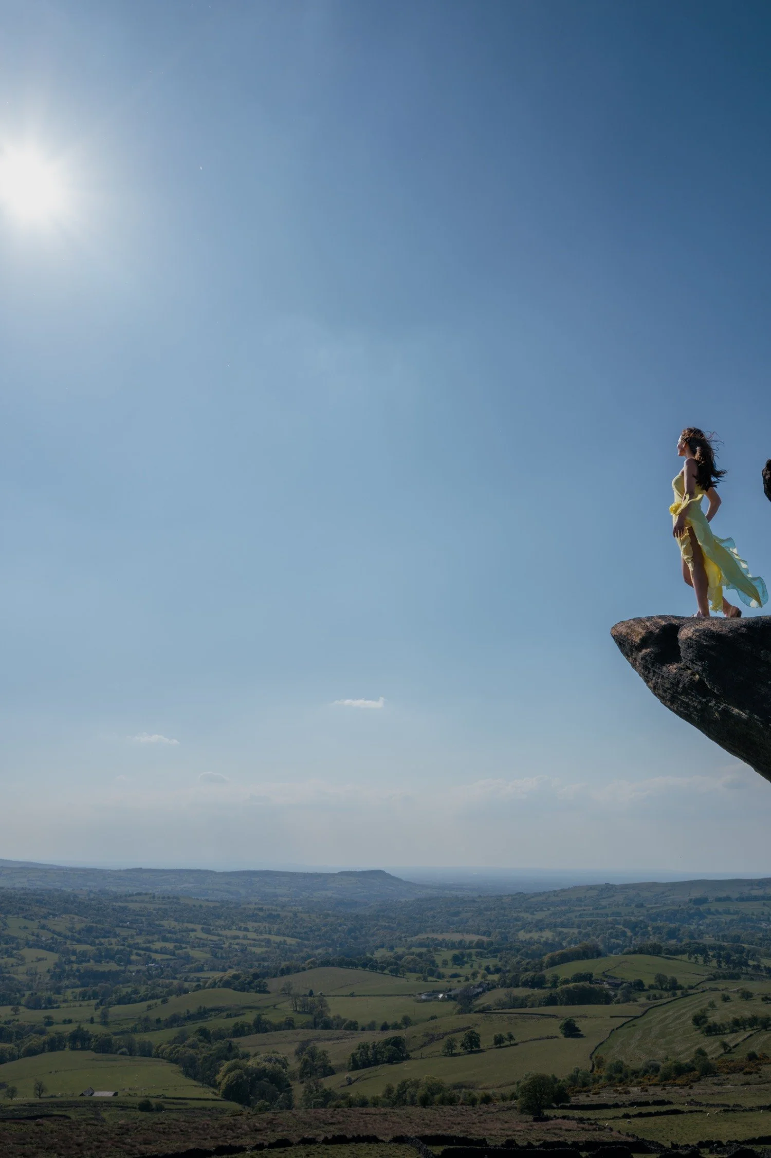 A woman in a yellow dress standing on the edge of a rock overlooking a vast landscape with rolling hills and a blue sky.