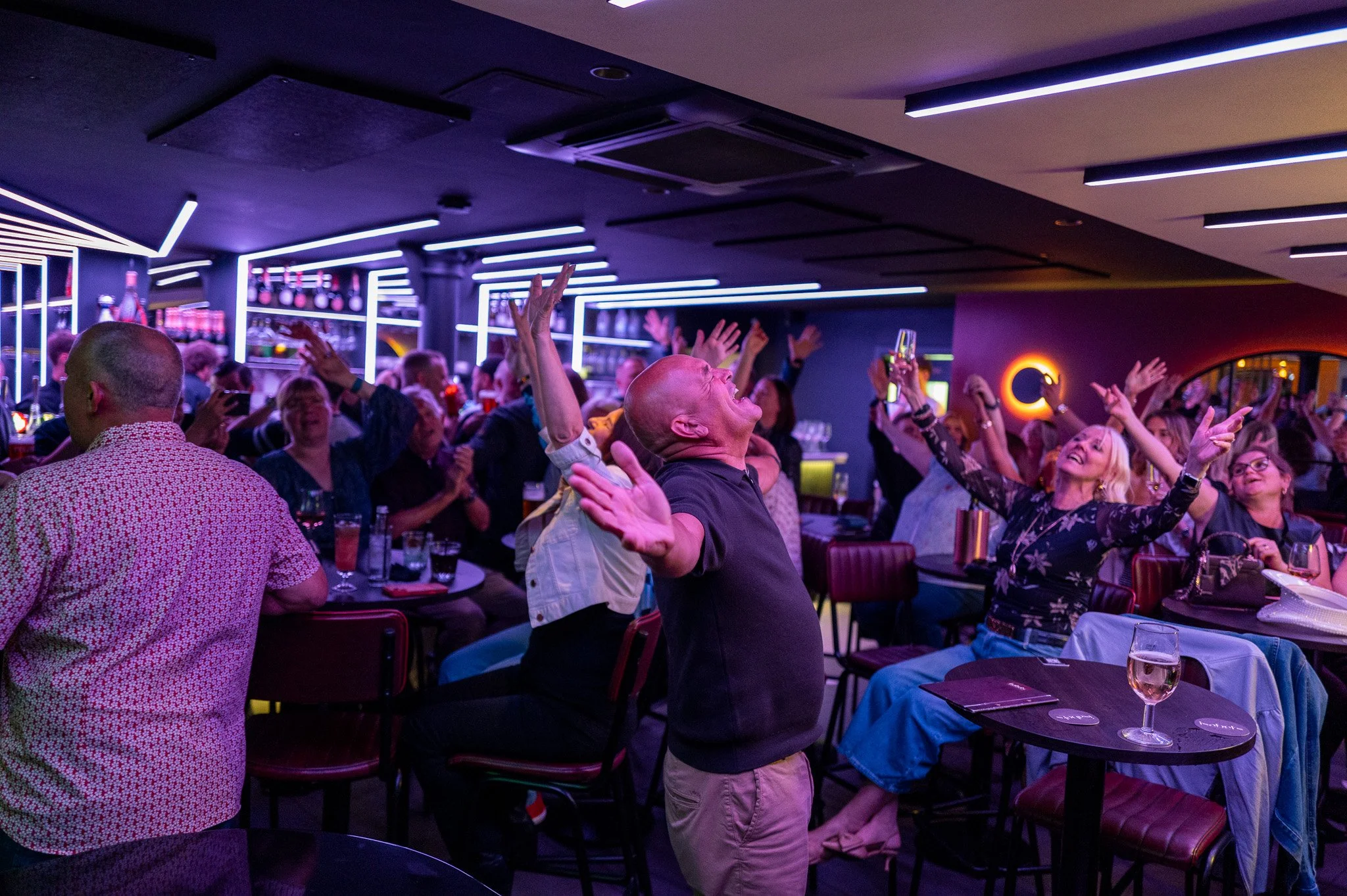 A lively nightclub scene filled with people dancing and raising their hands in the air. Purple and blue neon lighting glows across the room, creating a high-energy atmosphere. Tables with drinks surround the dance floor while guests smile, sing and c
