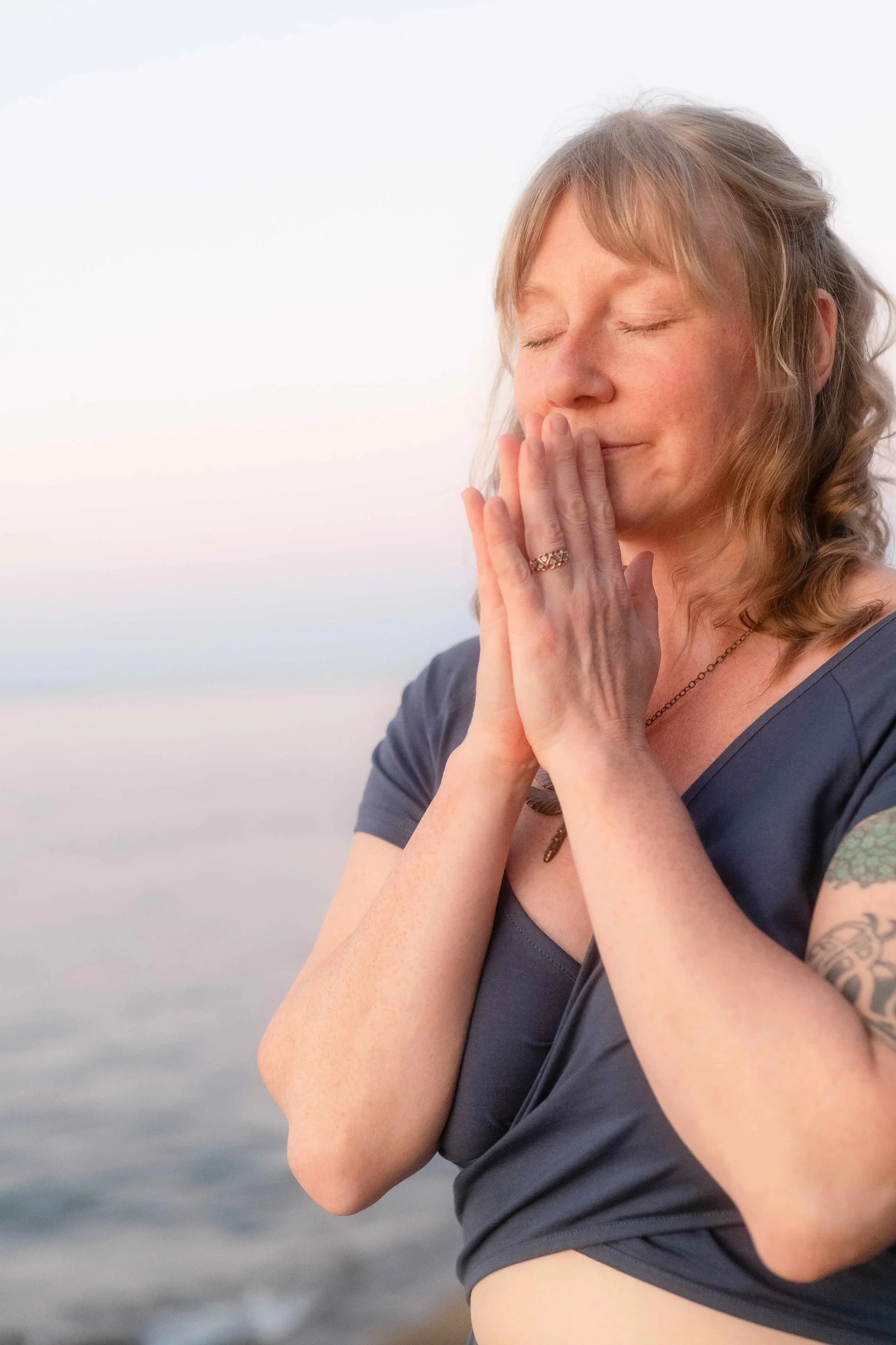 Juliana Christine with closed eyes and blonde hair, holding her hands in a prayer position near her face, standing outdoors with a blurred body of water in the background.