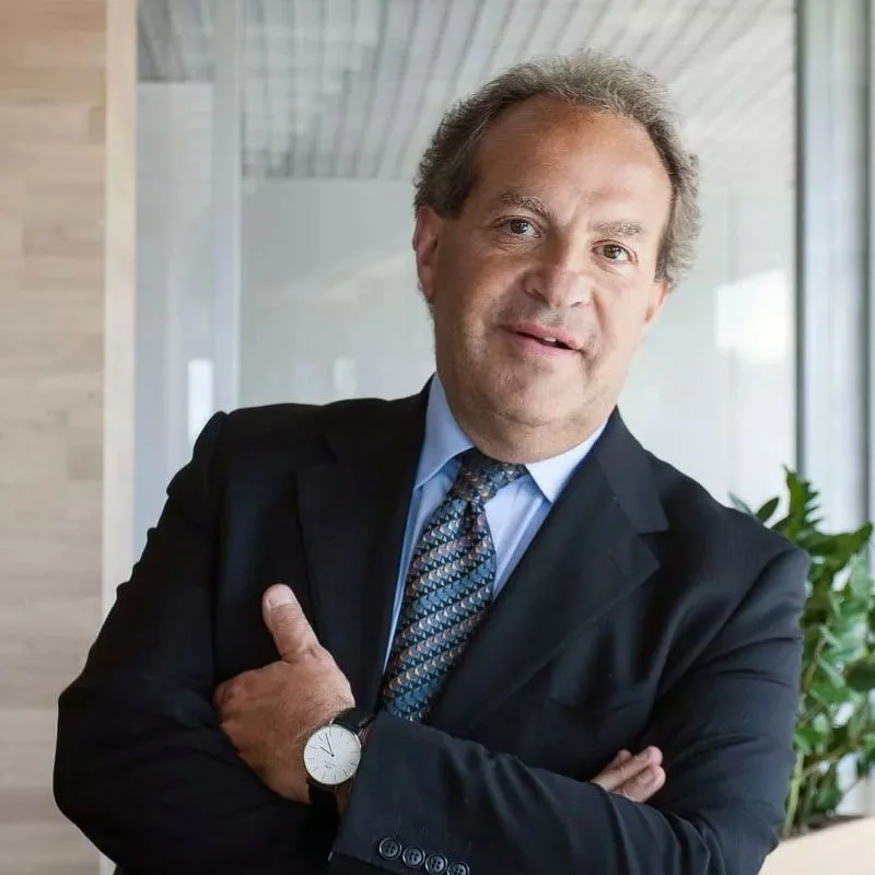 A middle-aged man with light brown hair and light skin, wearing a dark suit, light blue shirt, and patterned tie, smiling with arms crossed in an office setting, with large windows and green plants in the background.